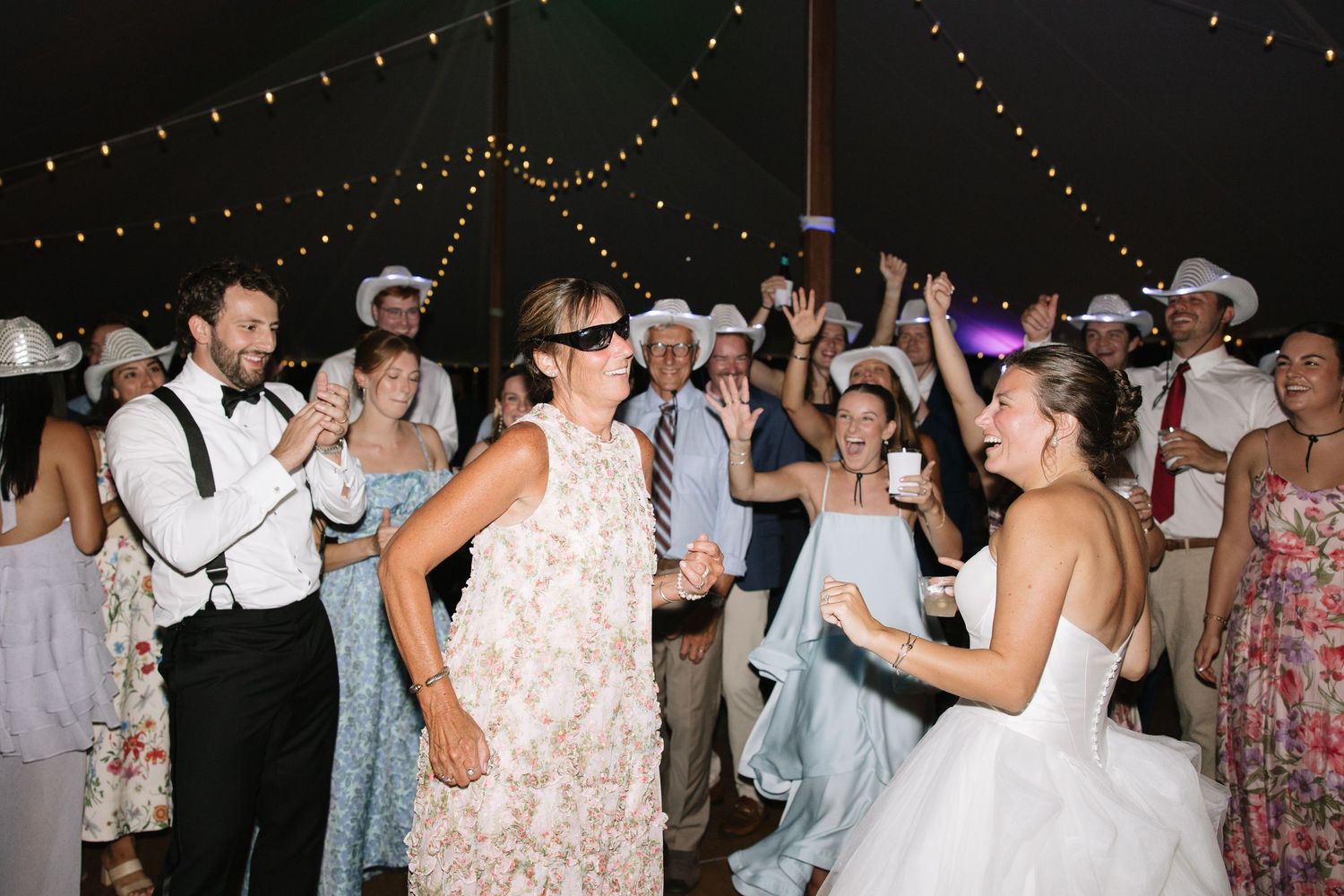 Bride and guests dance joyfully under string lights at lively outdoor wedding reception celebration.