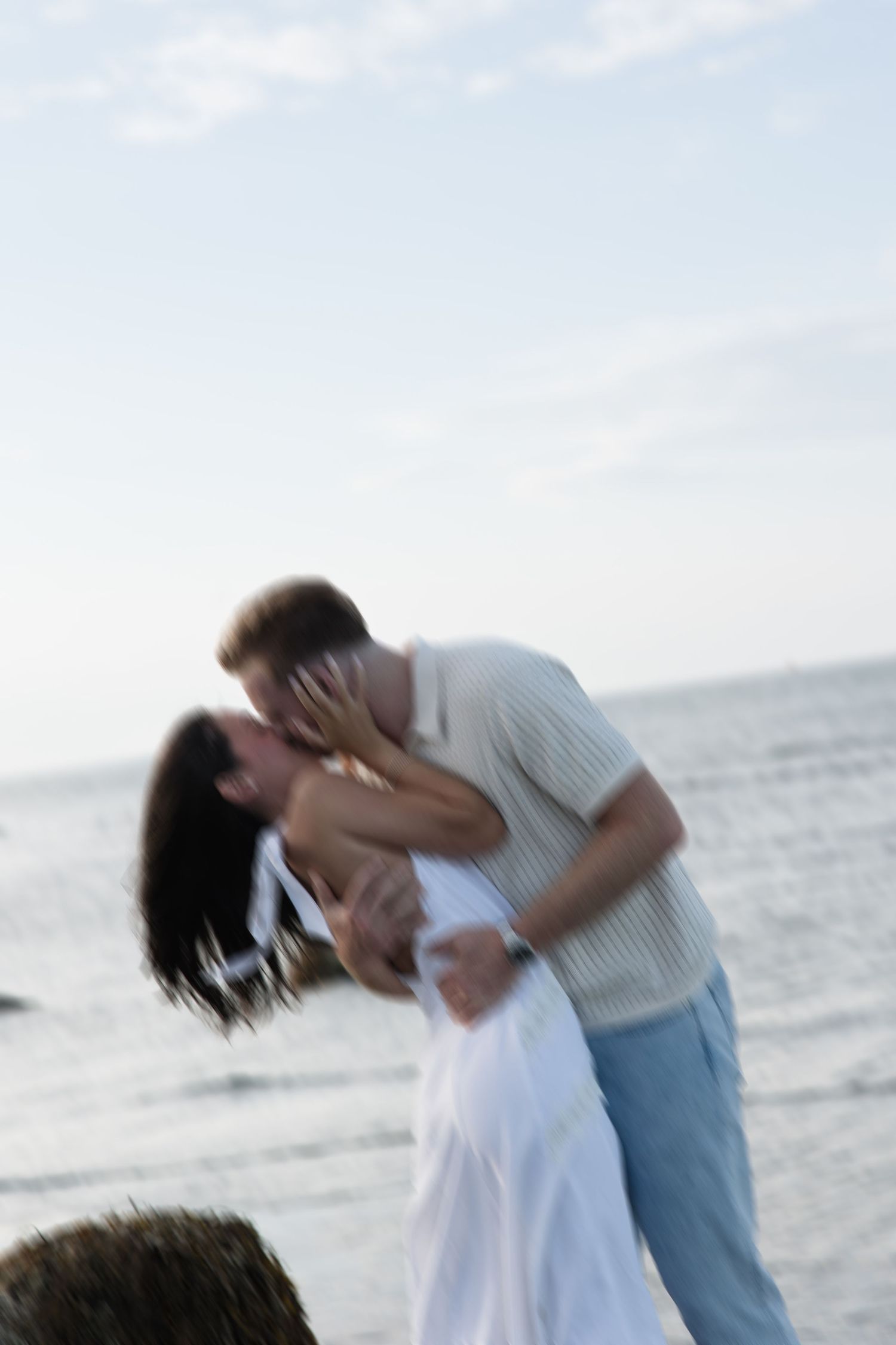Romantic couple sharing passionate kiss on rocky beach shoreline, woman in white dress, motion blur effect.