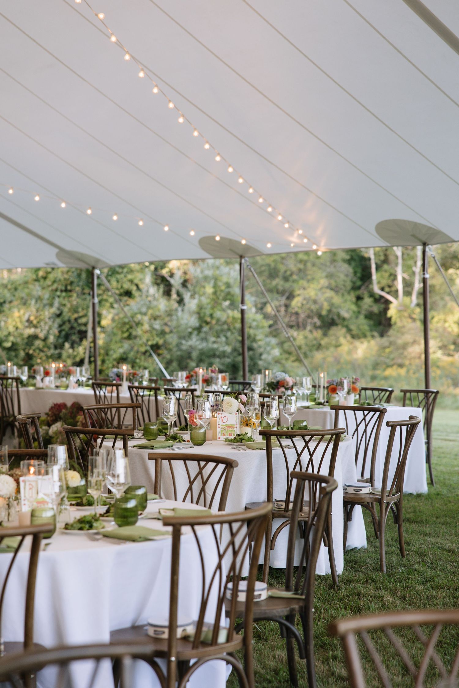 Elegant outdoor wedding reception under a white tent with string lights, round tables with white linens, and rustic wooden chairs.