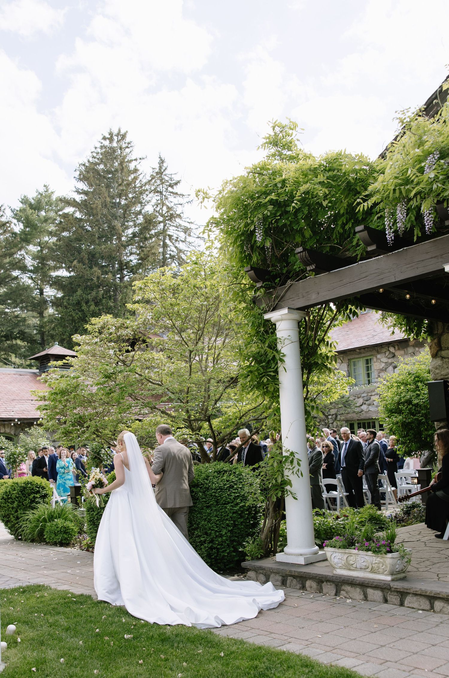 Bride in white gown and veil stands with escort at outdoor garden wedding ceremony beside stone building and pergola.