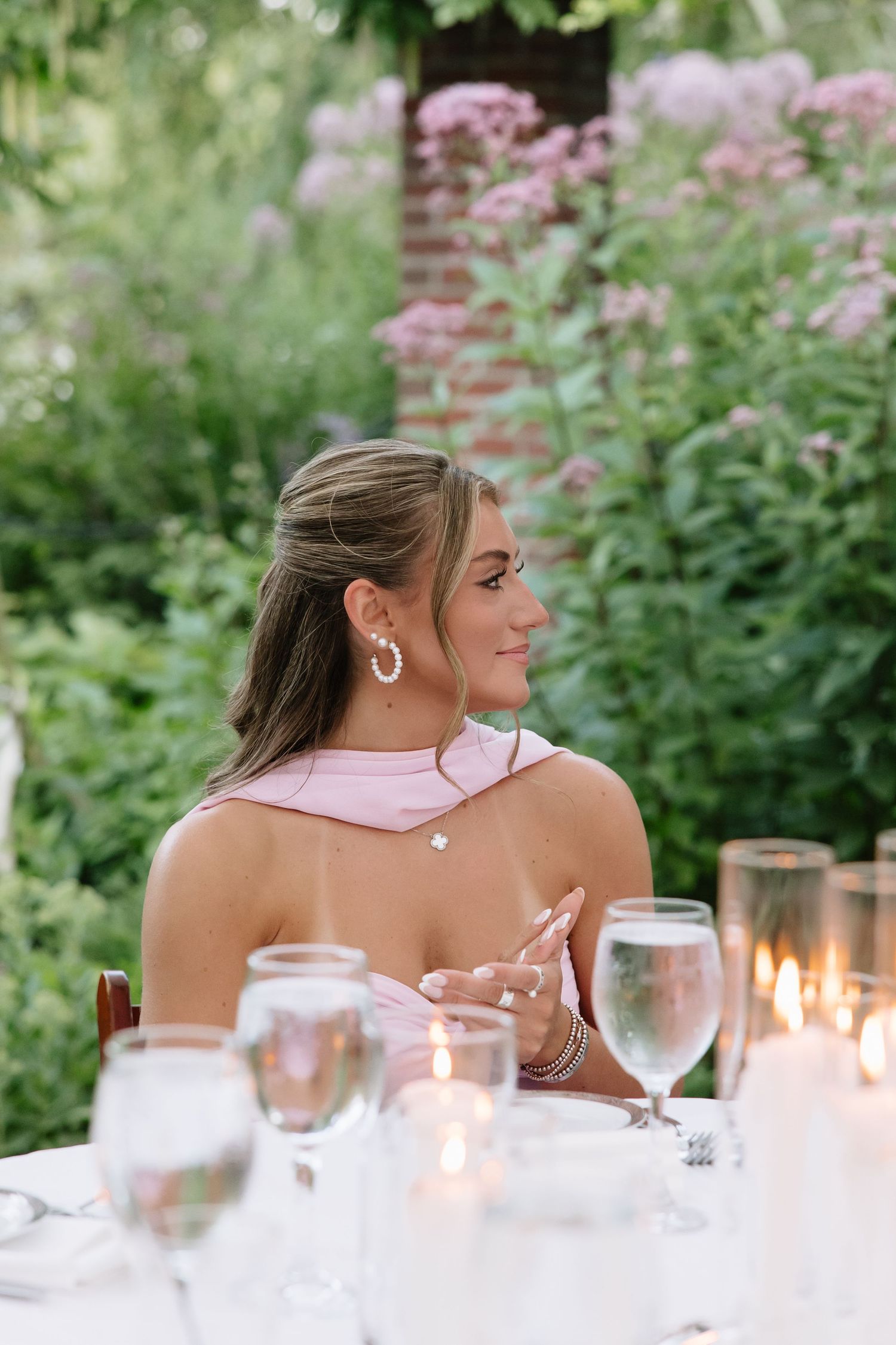 Woman in pink dress seated at elegant outdoor dinner table with candles and glassware, surrounded by lush green garden.