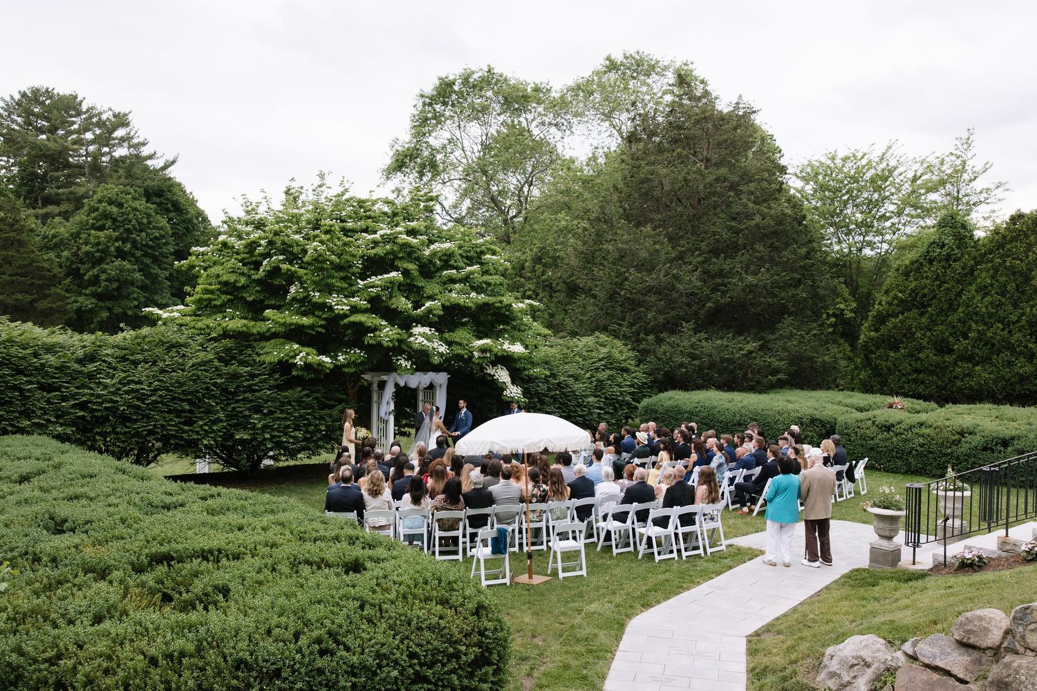 Outdoor wedding ceremony in a lush green garden with white chairs, floral arch, and guests gathered on a cloudy day.
