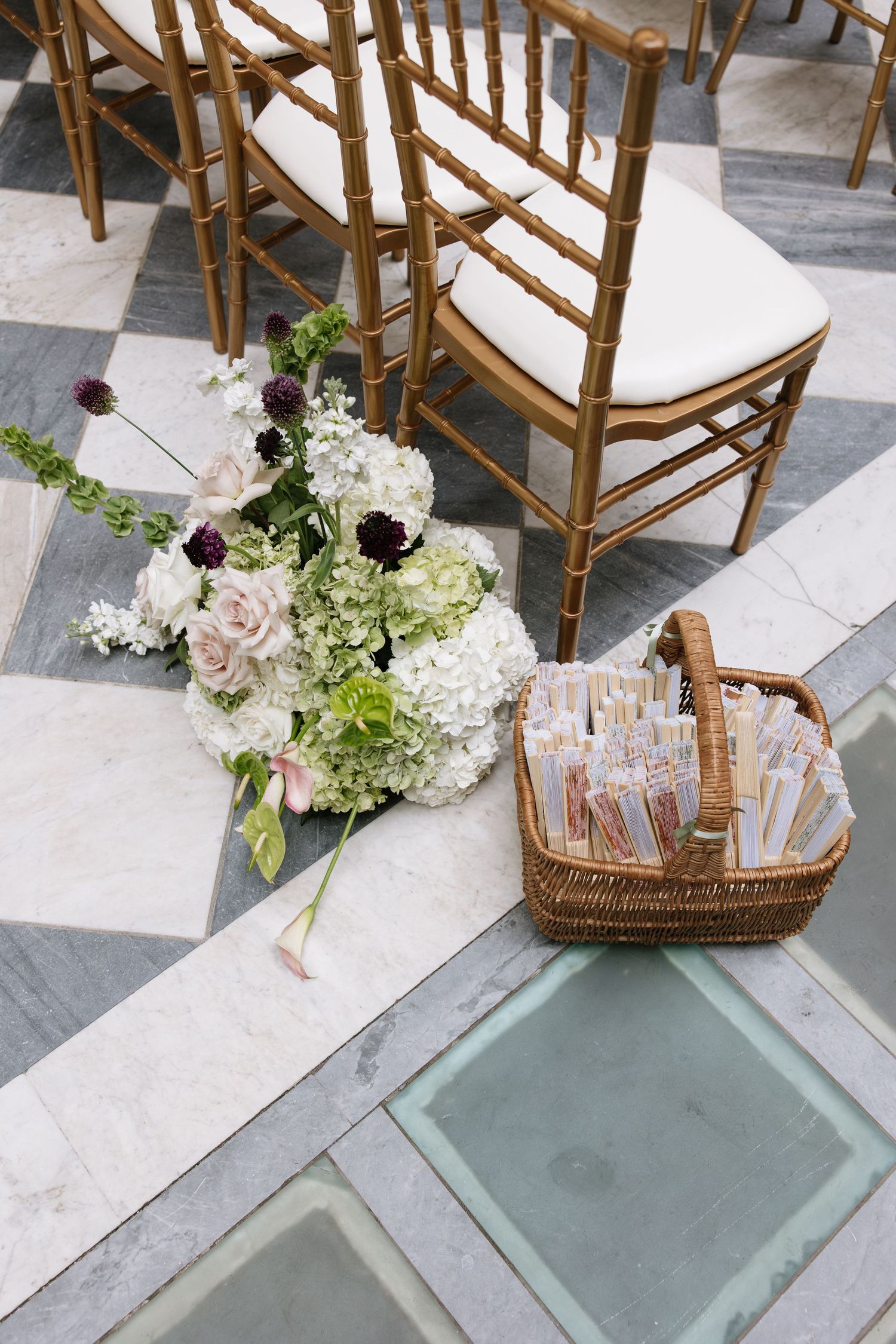 Elegant wedding ceremony aisle with gold chiavari chairs, white floral arrangement, and wicker basket of programs on marble floor.