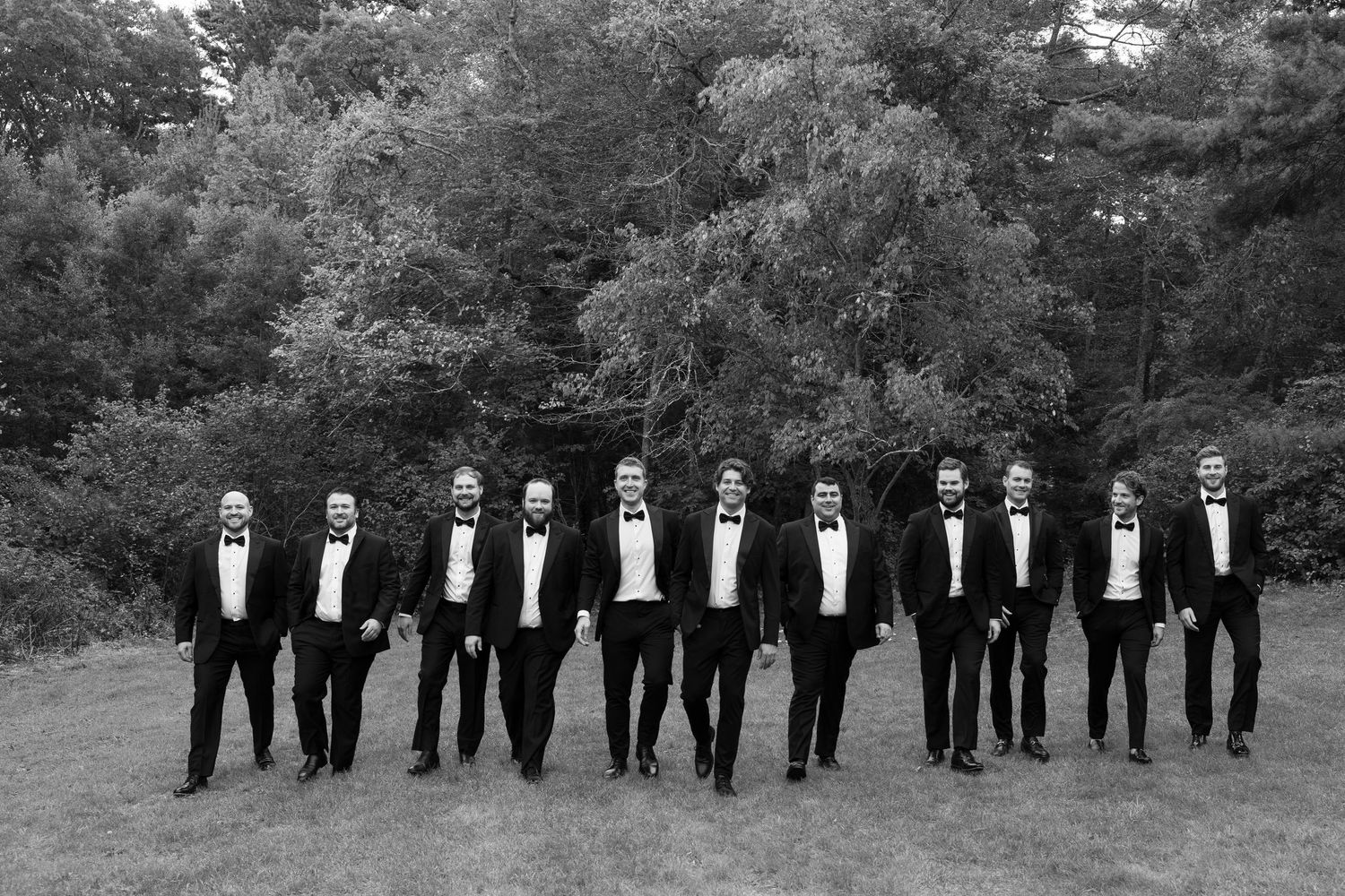 Black and white panoramic shot of groomsmen lined up in tuxedos outdoors.