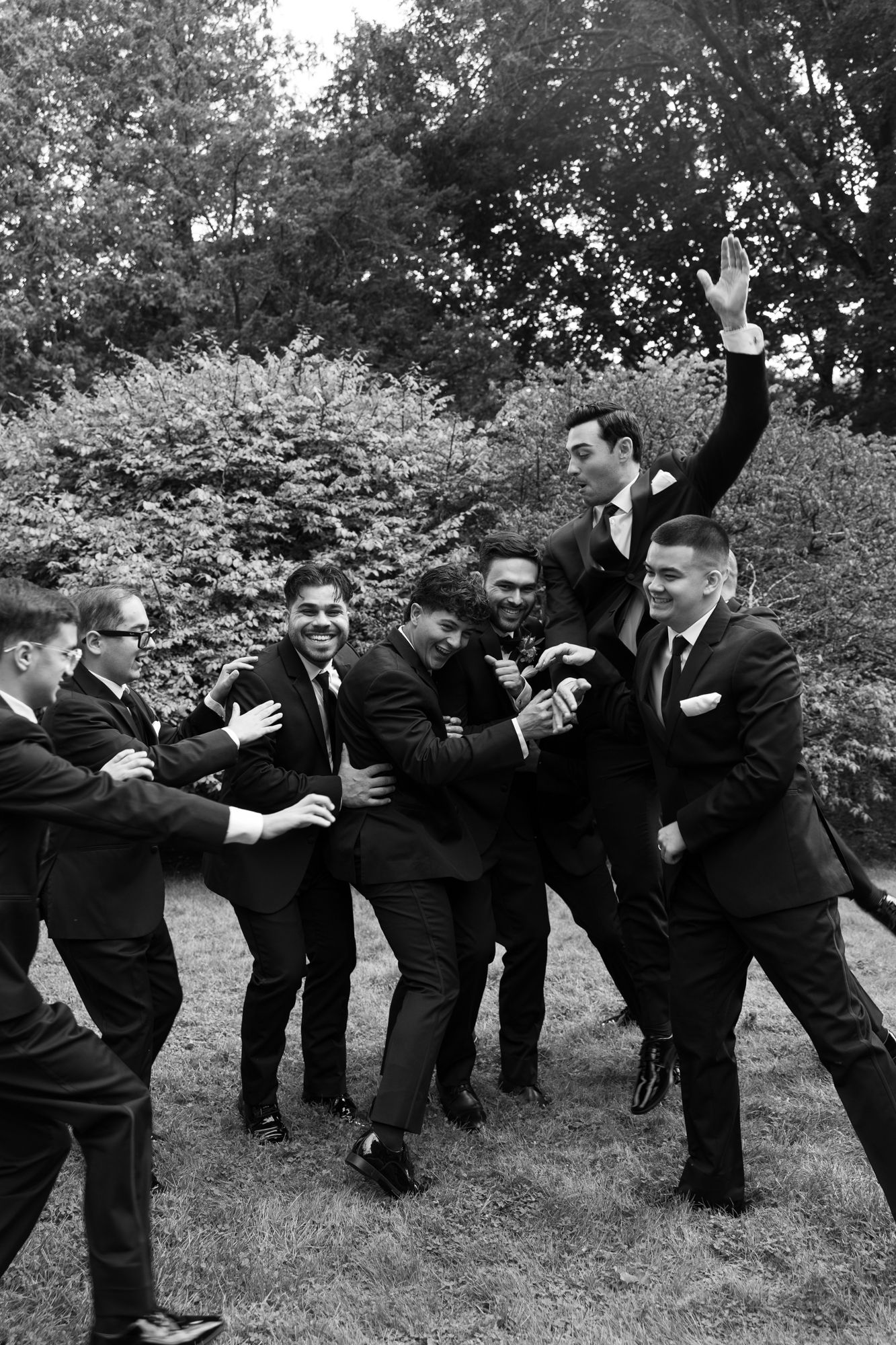Groomsmen performing a fun synchronized pose in black suits on grassy terrain.