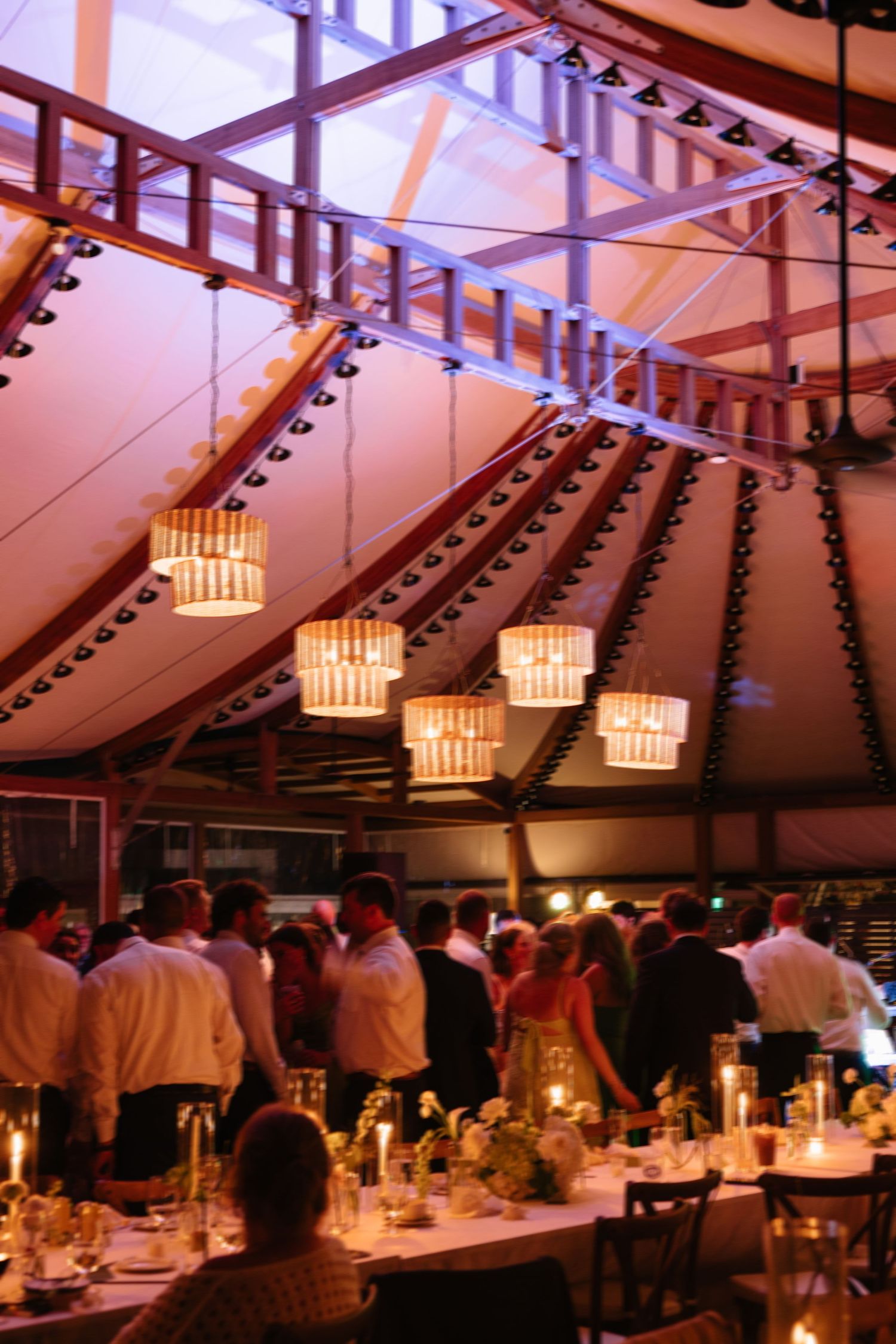 Romantic wedding reception under a dramatic tent ceiling with warm lighting, chandelier pendants, and guests dancing at night.