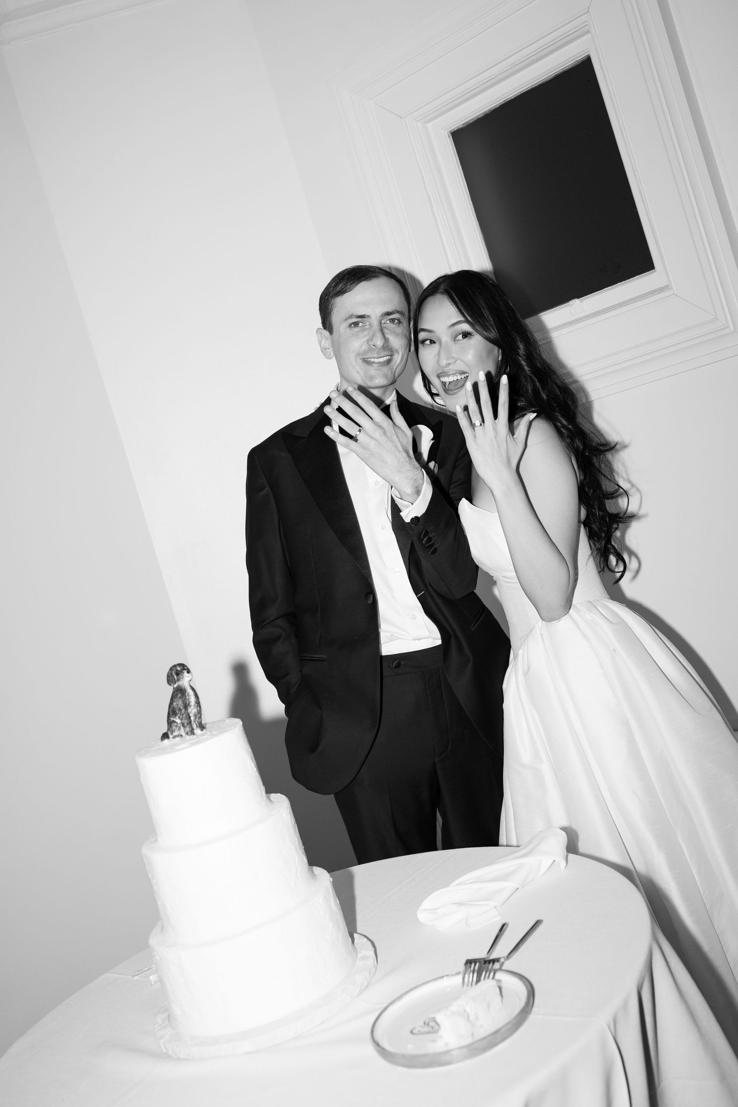 Black and white photo of newlyweds showing wedding rings beside tiered cake.