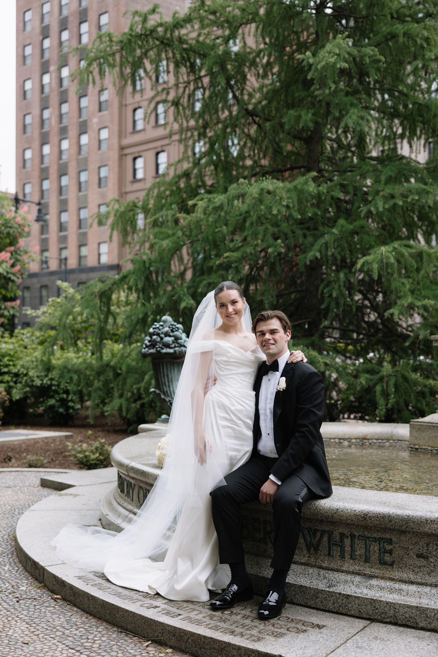 Bride and groom posing at a stone fountain in a lush urban garden on their wedding day.