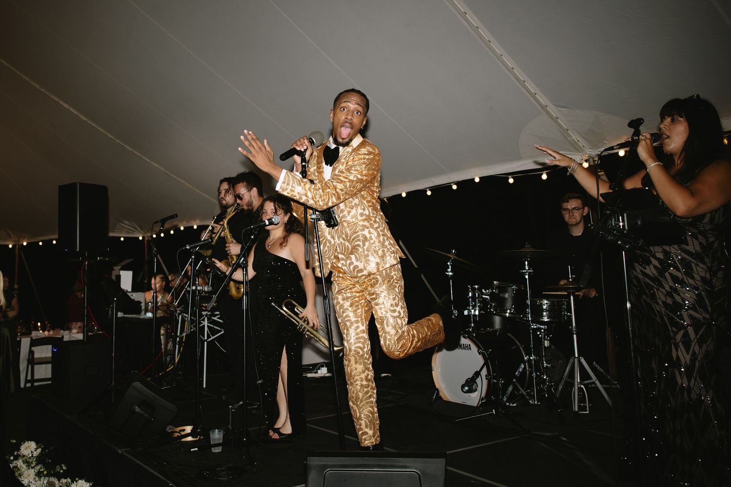 Performer in gold sequin suit singing on stage at elegant tented wedding reception with live band.