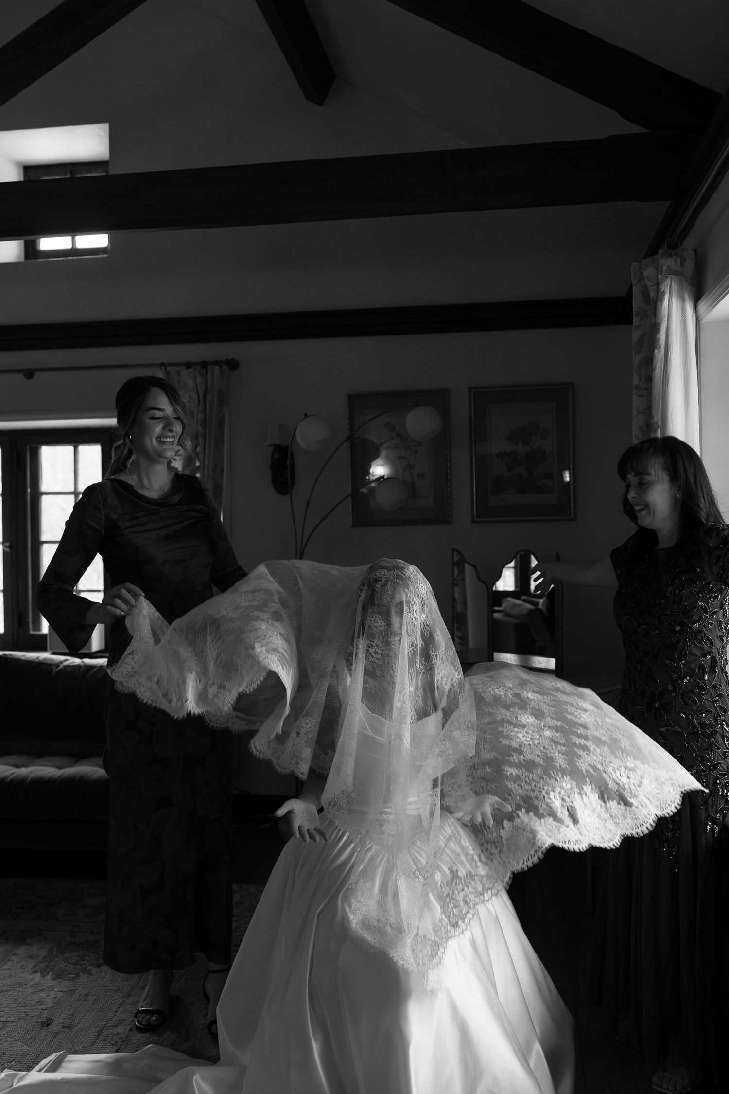 Black and white photo of bride having veil placed by two women assistants in elegant indoor setting.