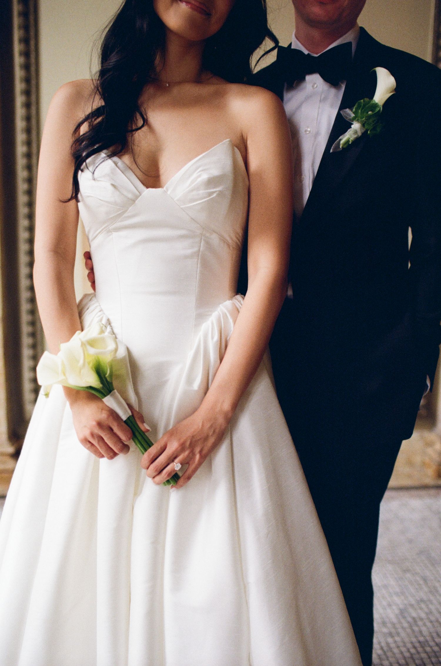 Bride in strapless white ball gown holding calla lily bouquet stands with groom in black tuxedo with bow tie.