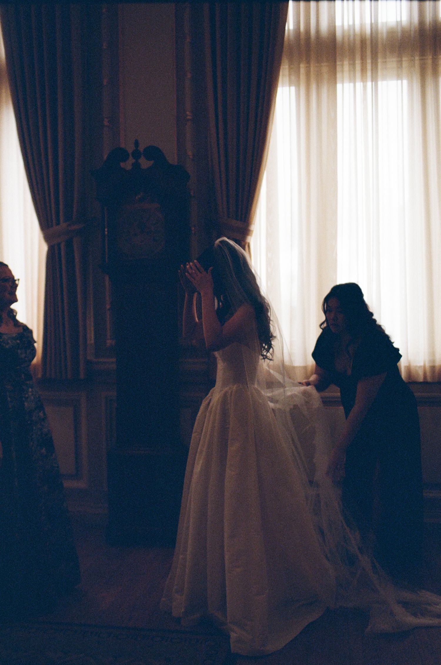 Bride in white wedding gown praying with veil, flanked by attendants near grandfather clock by sheer curtains.