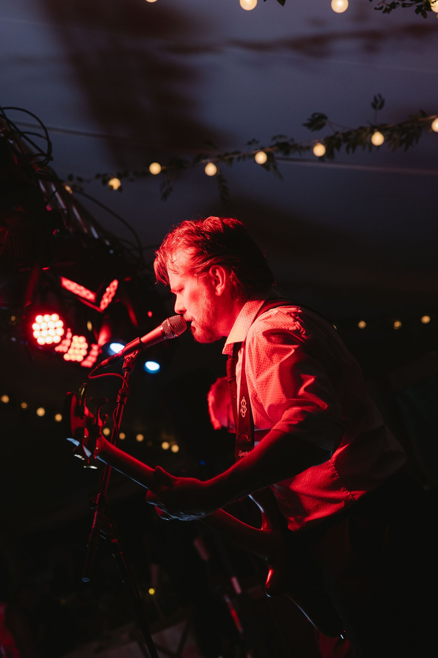 Male musician performing on stage under red lights and string lights at an outdoor night concert.