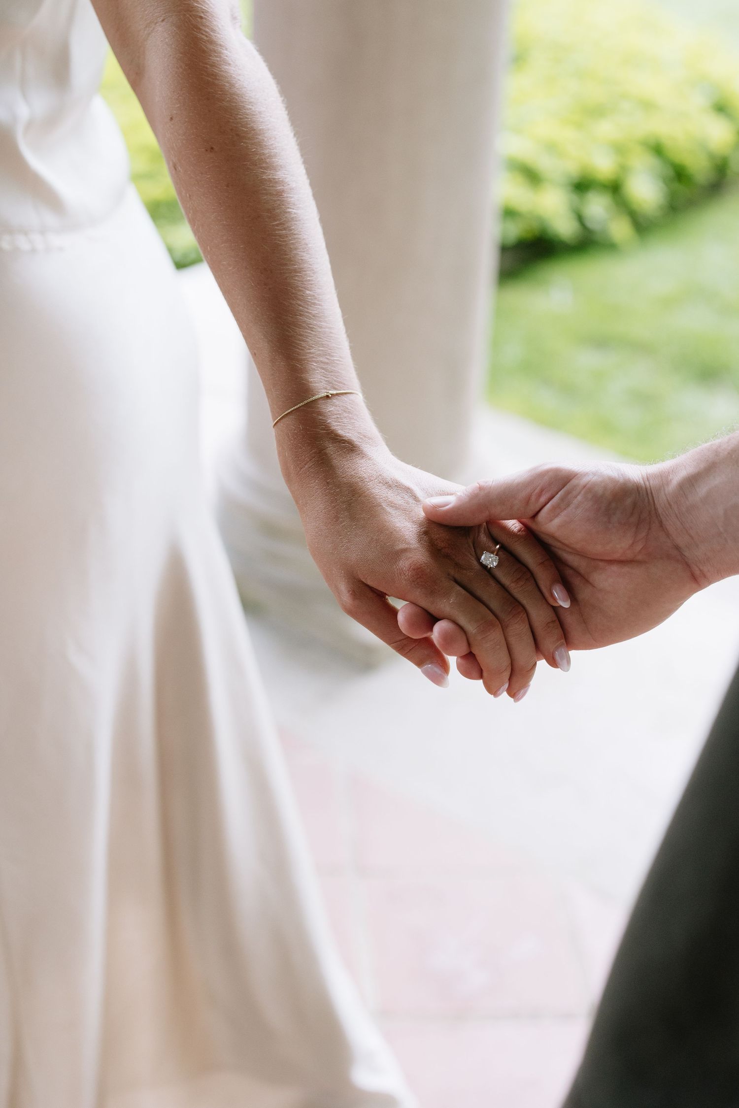 Bride in white dress holding hands with groom, showcasing elegant diamond engagement ring and gold bracelet at wedding.