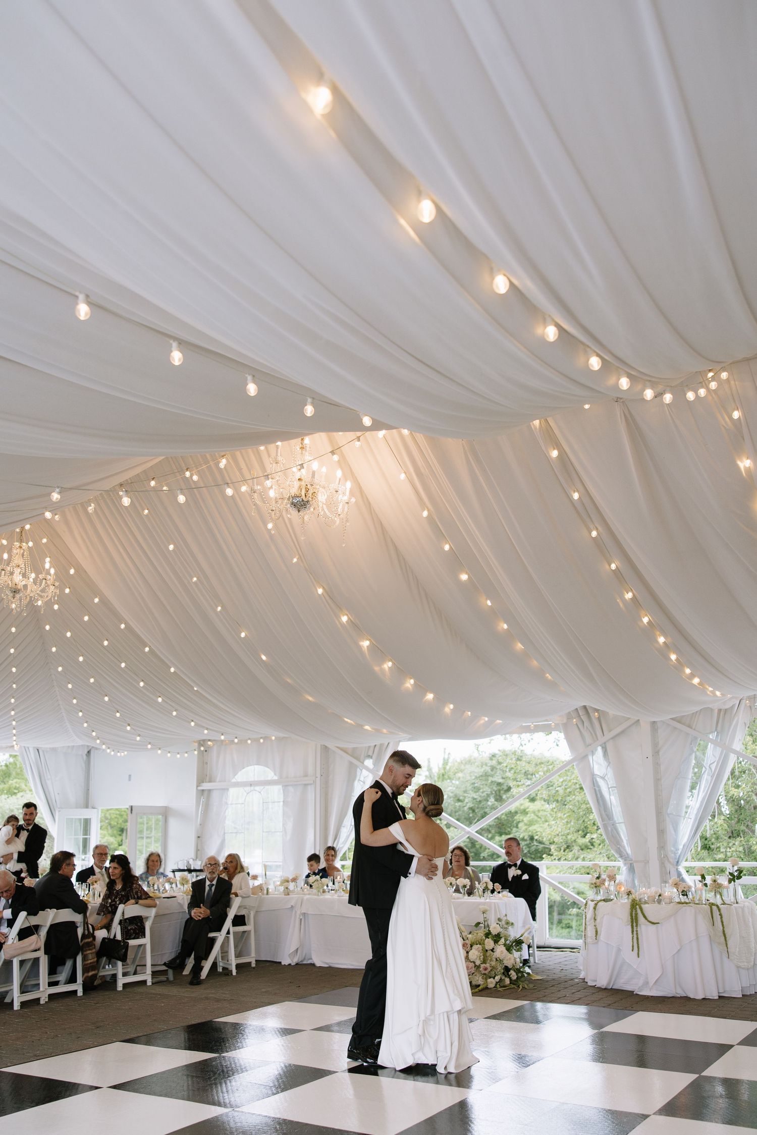 Wedding reception dance floor under white tent with string lights and dancing guests.
