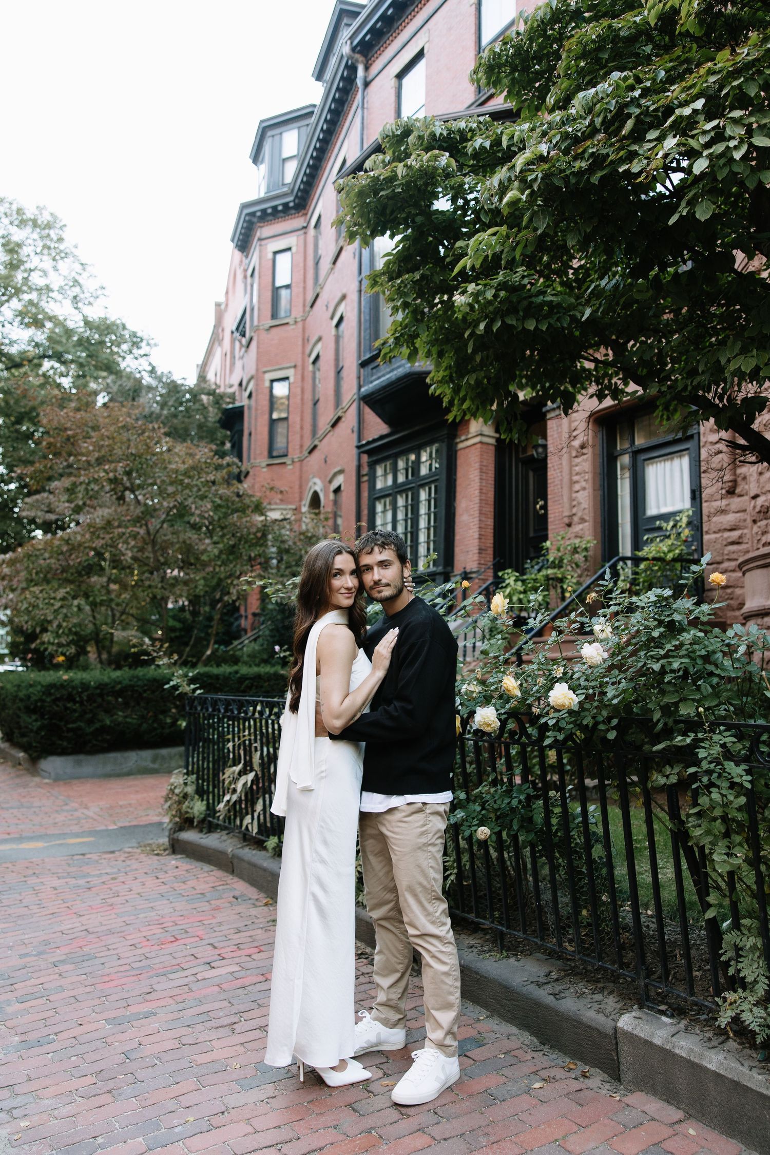 Couple in casual formal wear posing in front of historic brownstone building.