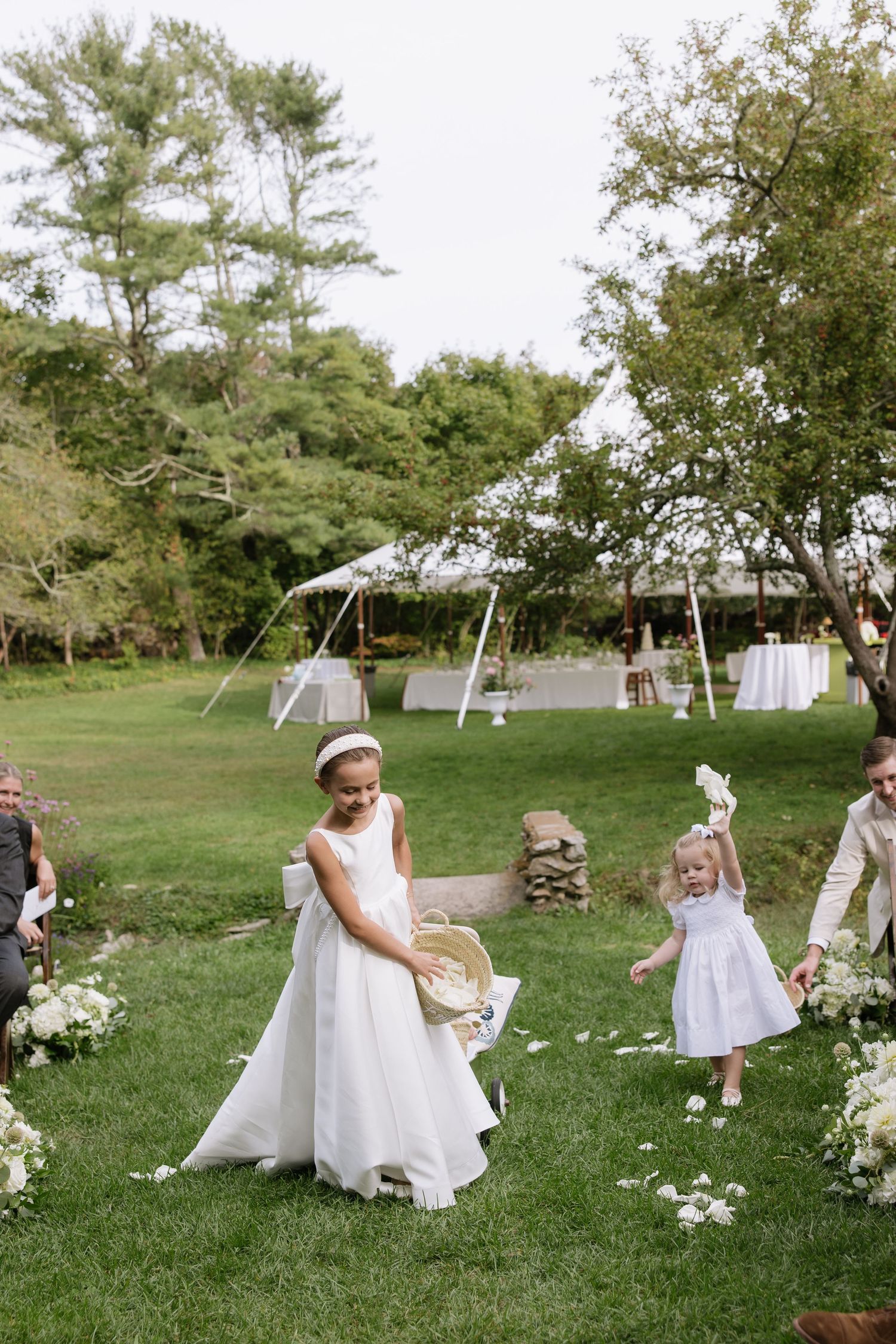 Young attendants in white dresses play on a lawn with outdoor reception setup in background.