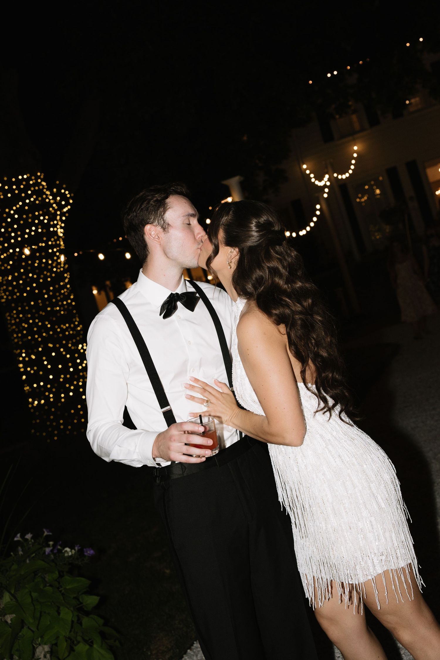 Couple sharing a romantic kiss at night, man in white shirt with suspenders and bow tie, woman in white fringe dress.