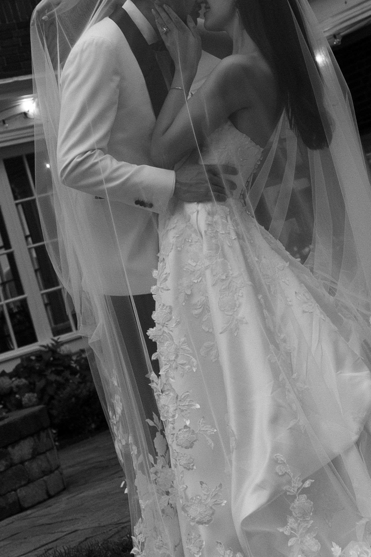 Black and white wedding photo of bride in floral lace gown and veil embracing groom in white suit outdoors at night.