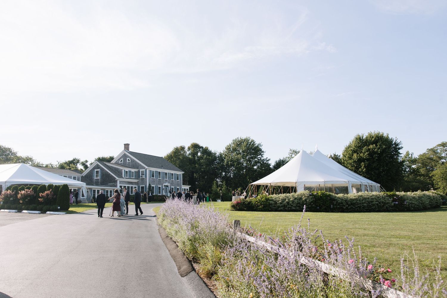 Elegant outdoor wedding venue with white reception tents on manicured lawn beside a classic gray shingle-style estate.