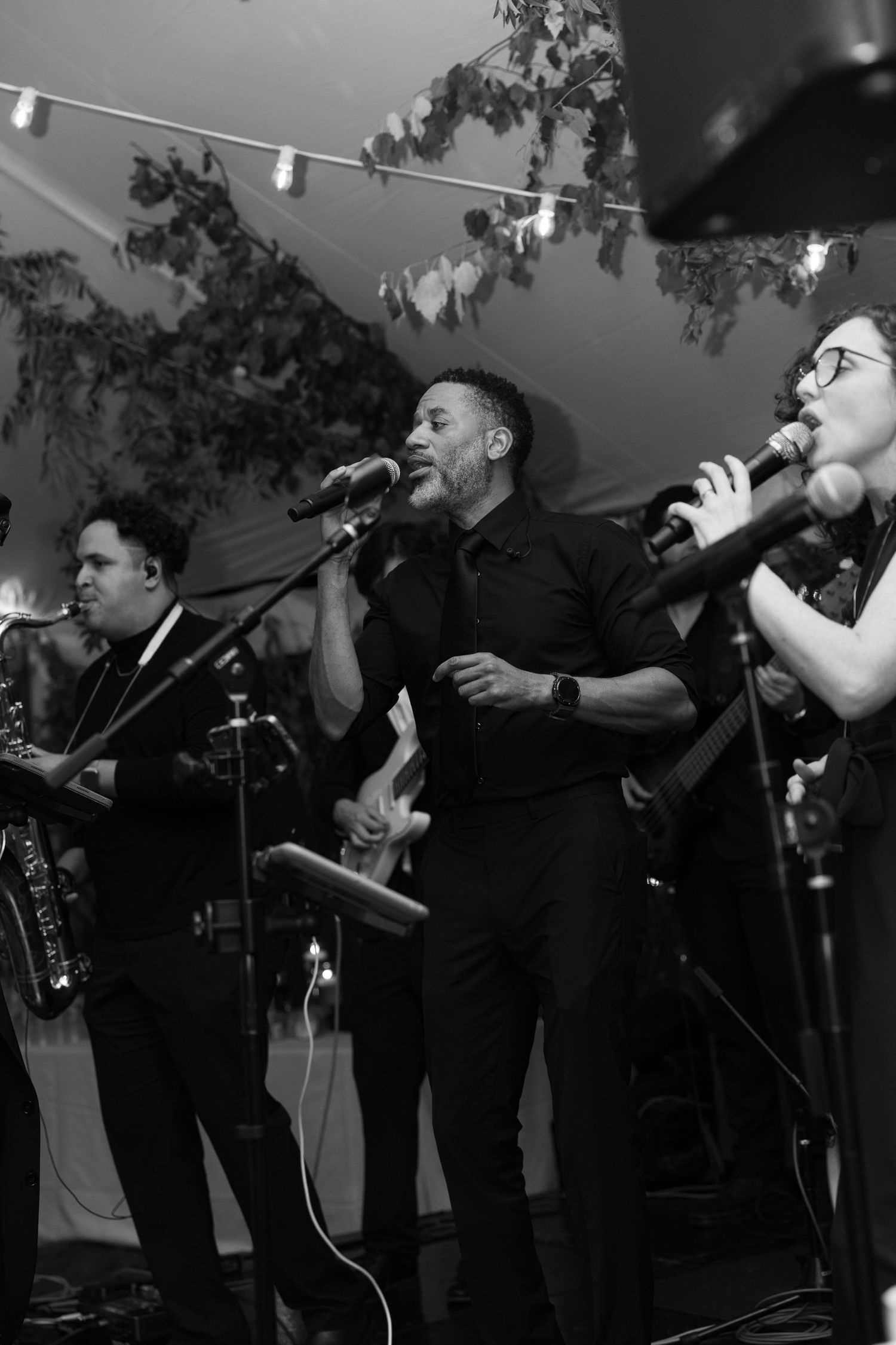 Black and white photo of a live band performing, two singers at microphones in all-black attire at an elegant event.