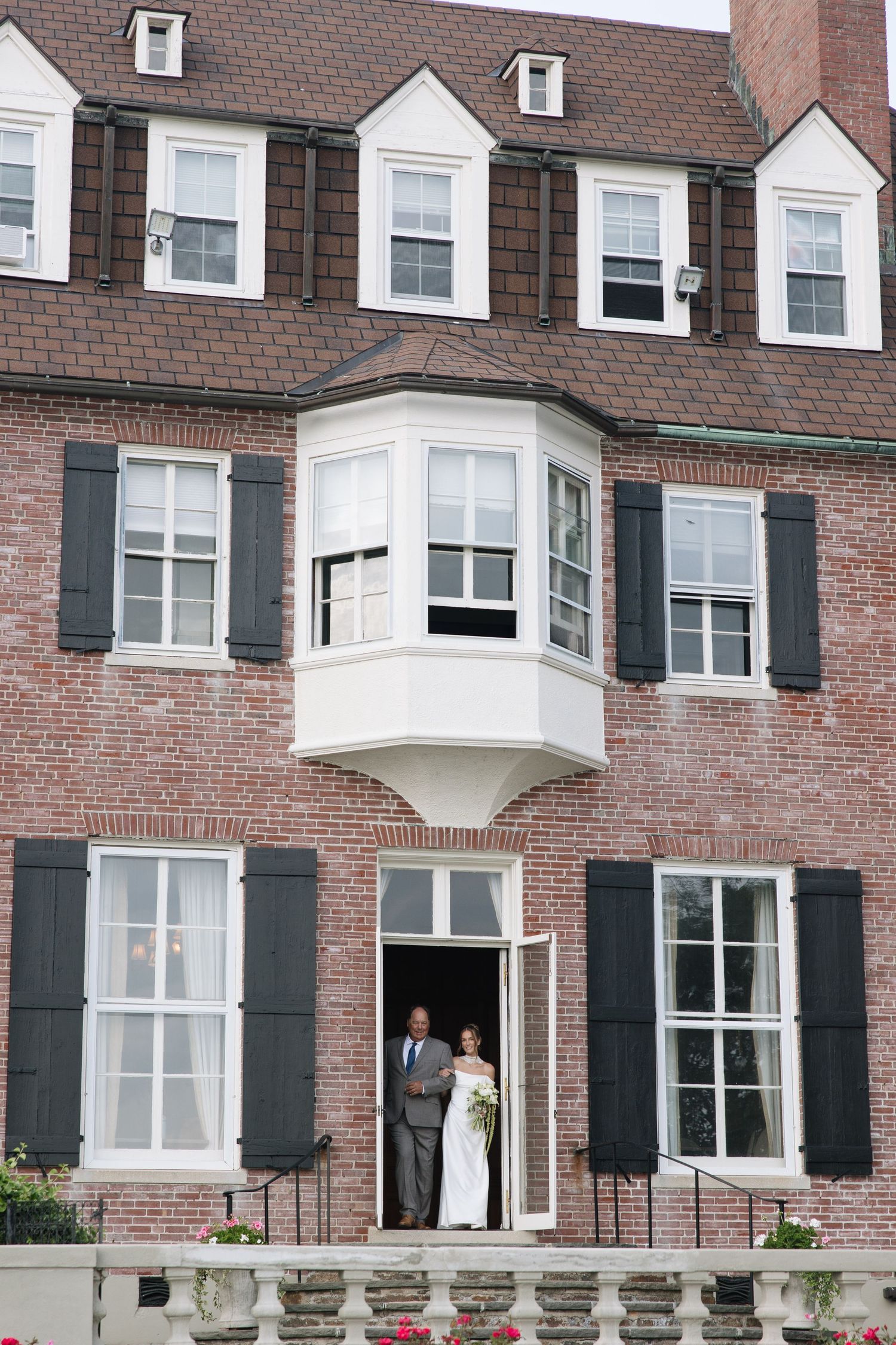 Bride and groom stand in doorway of brick mansion with dark shutters during their wedding day.