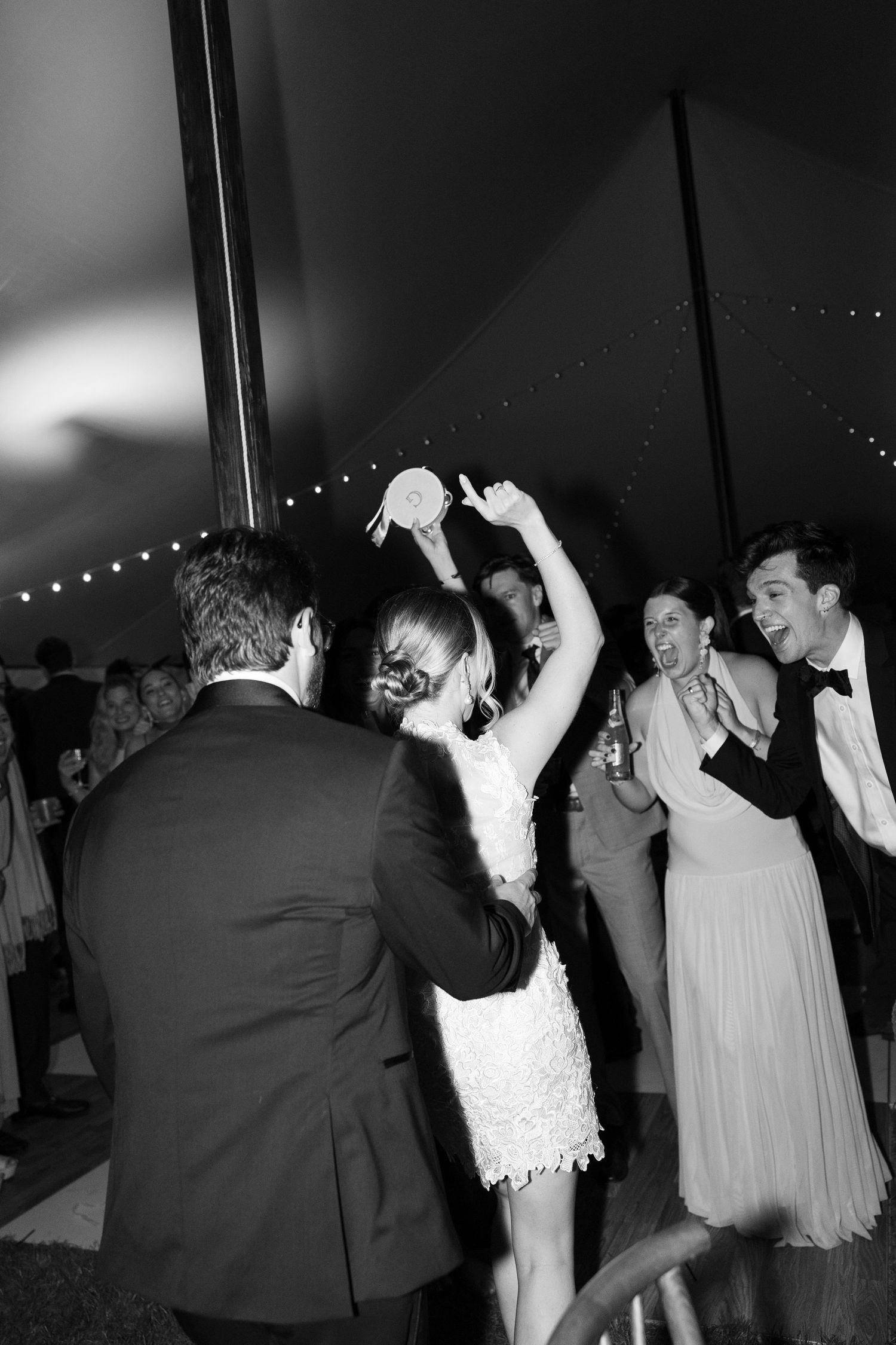 Black and white photo of wedding guests dancing joyfully under a tent at a lively evening reception.