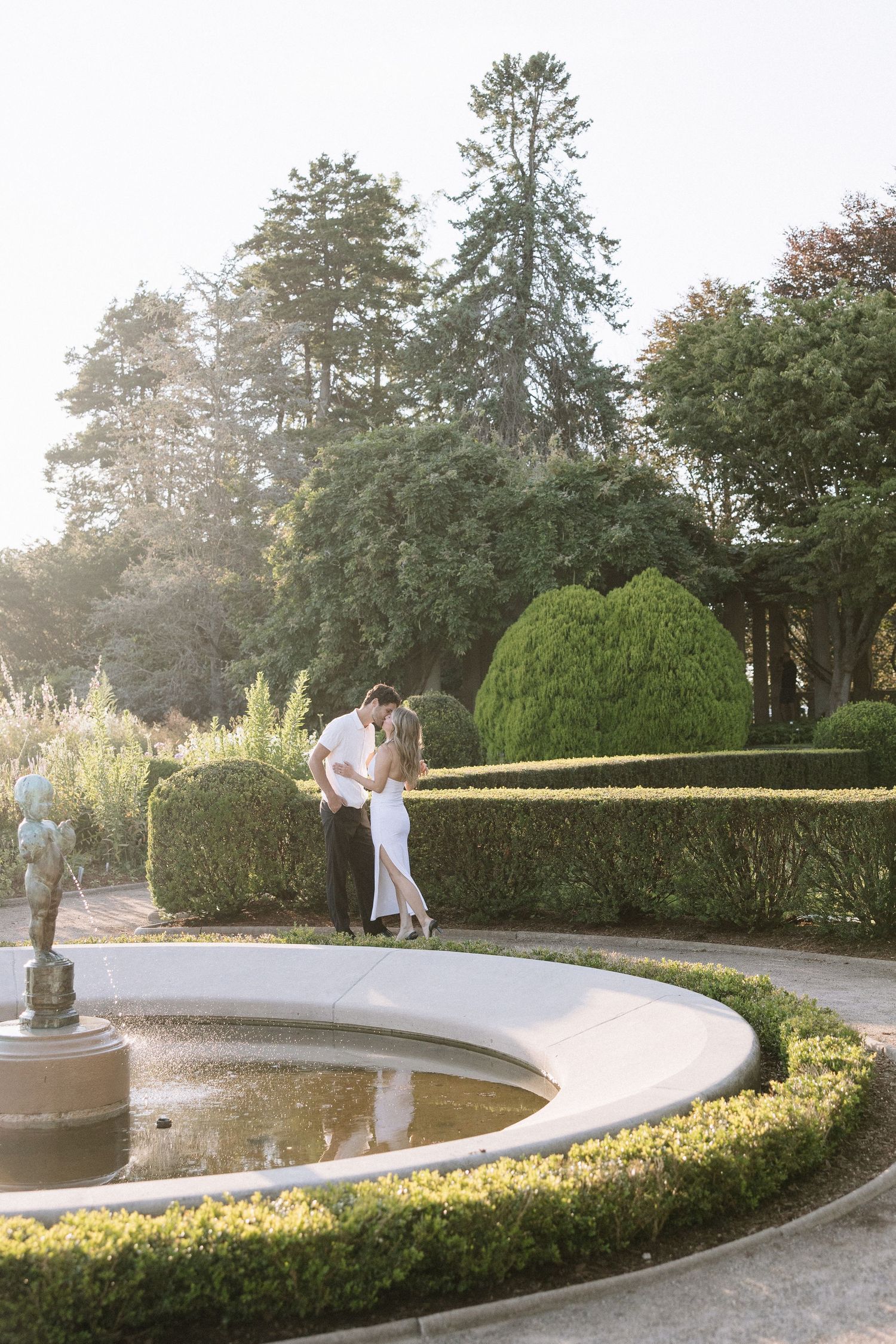 Couple embracing in white attire beside a circular garden fountain surrounded by manicured hedges and tall trees.