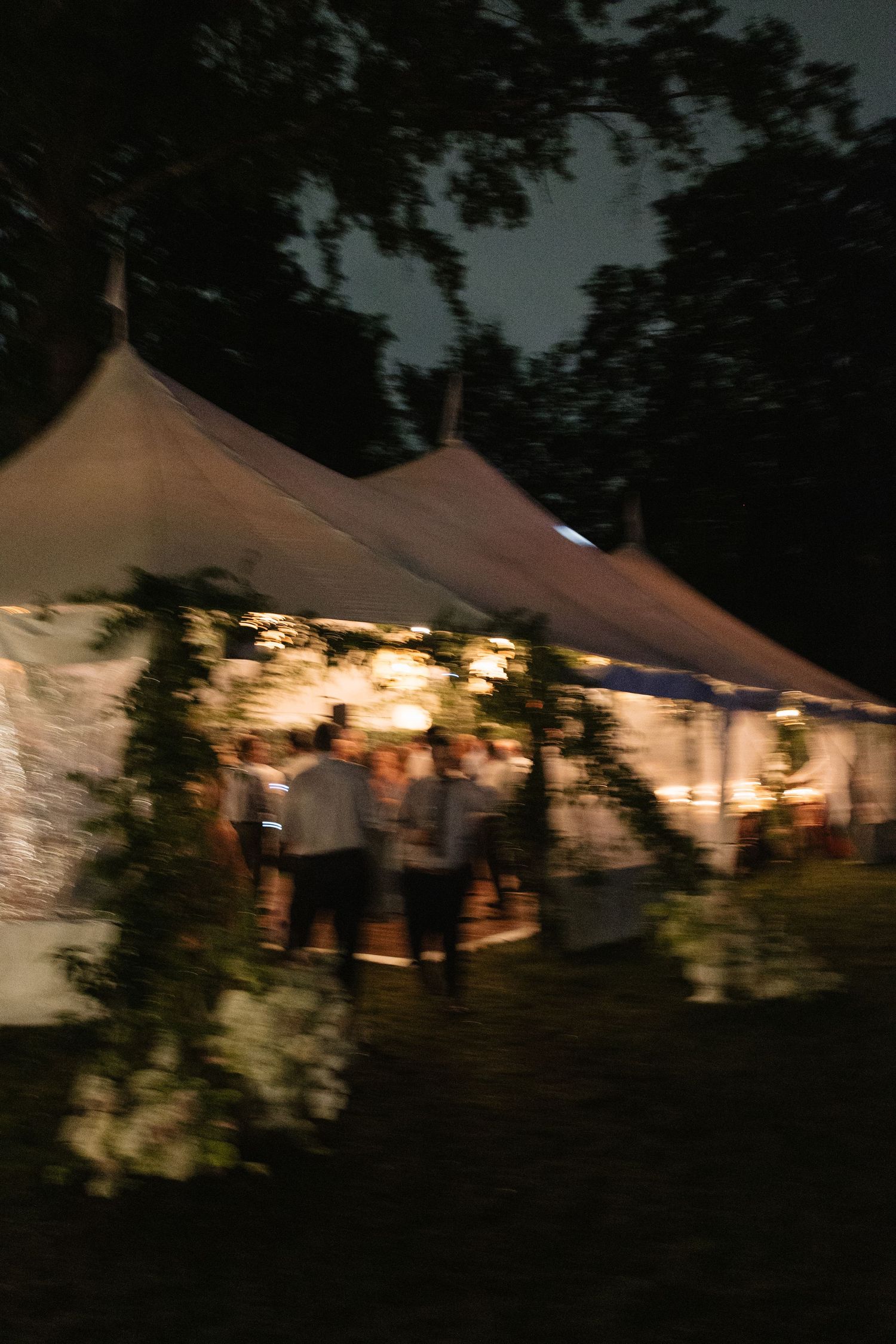 Illuminated white wedding tent at night surrounded by lush greenery with guests gathered outside during an outdoor reception.