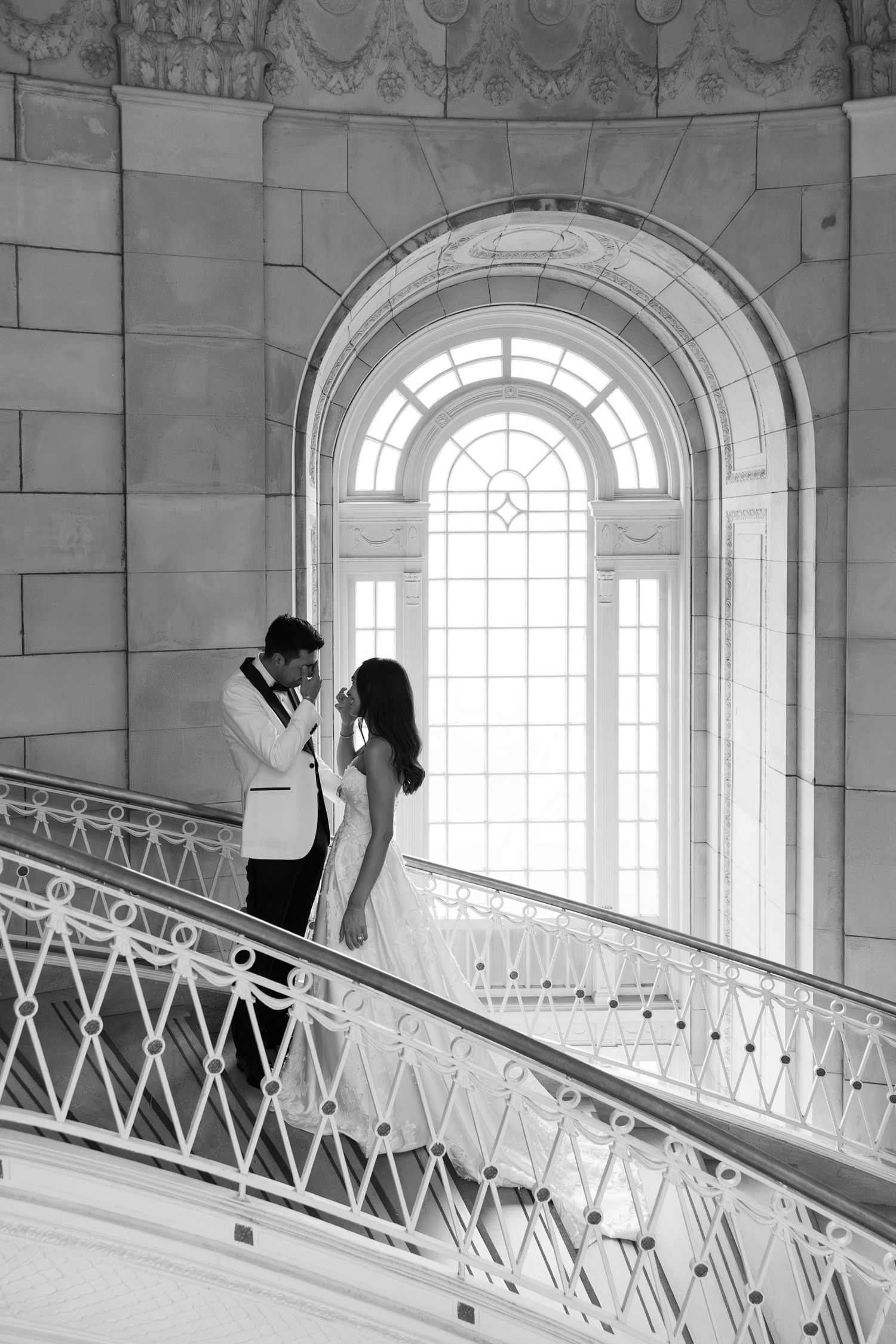 Black and white photo of a bride and groom embracing on an elegant staircase with arched window backdrop.
