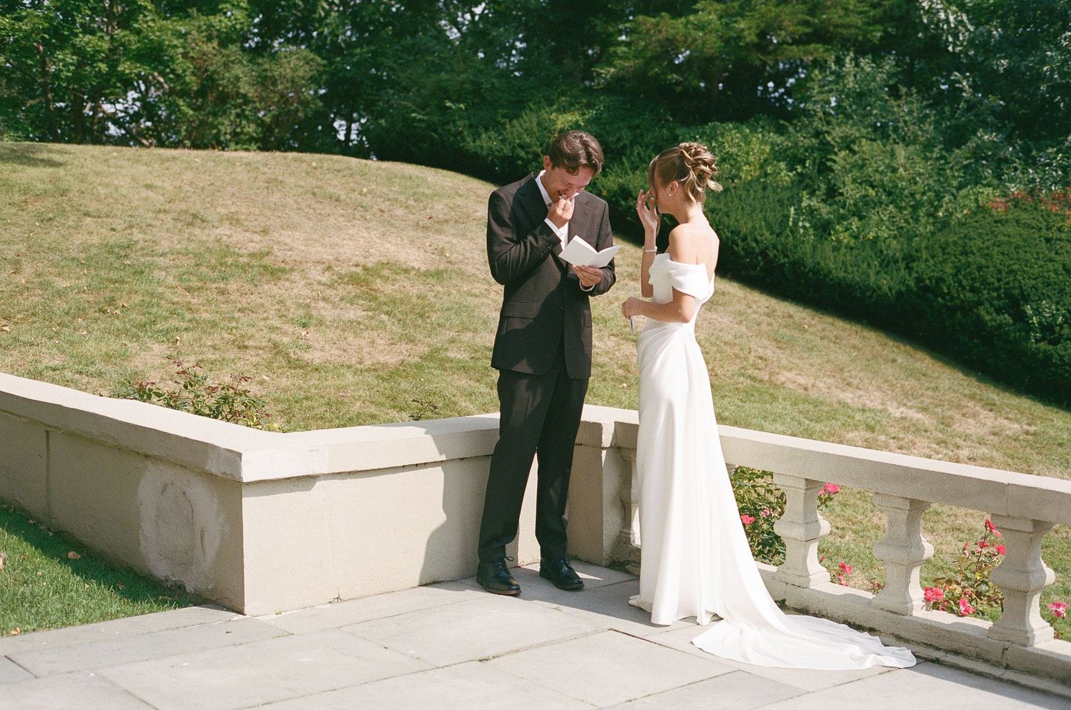 Groom in a black suit wipes his eyes while standing beside a bride in an off-shoulder white gown on a stone terrace with a grassy hillside.