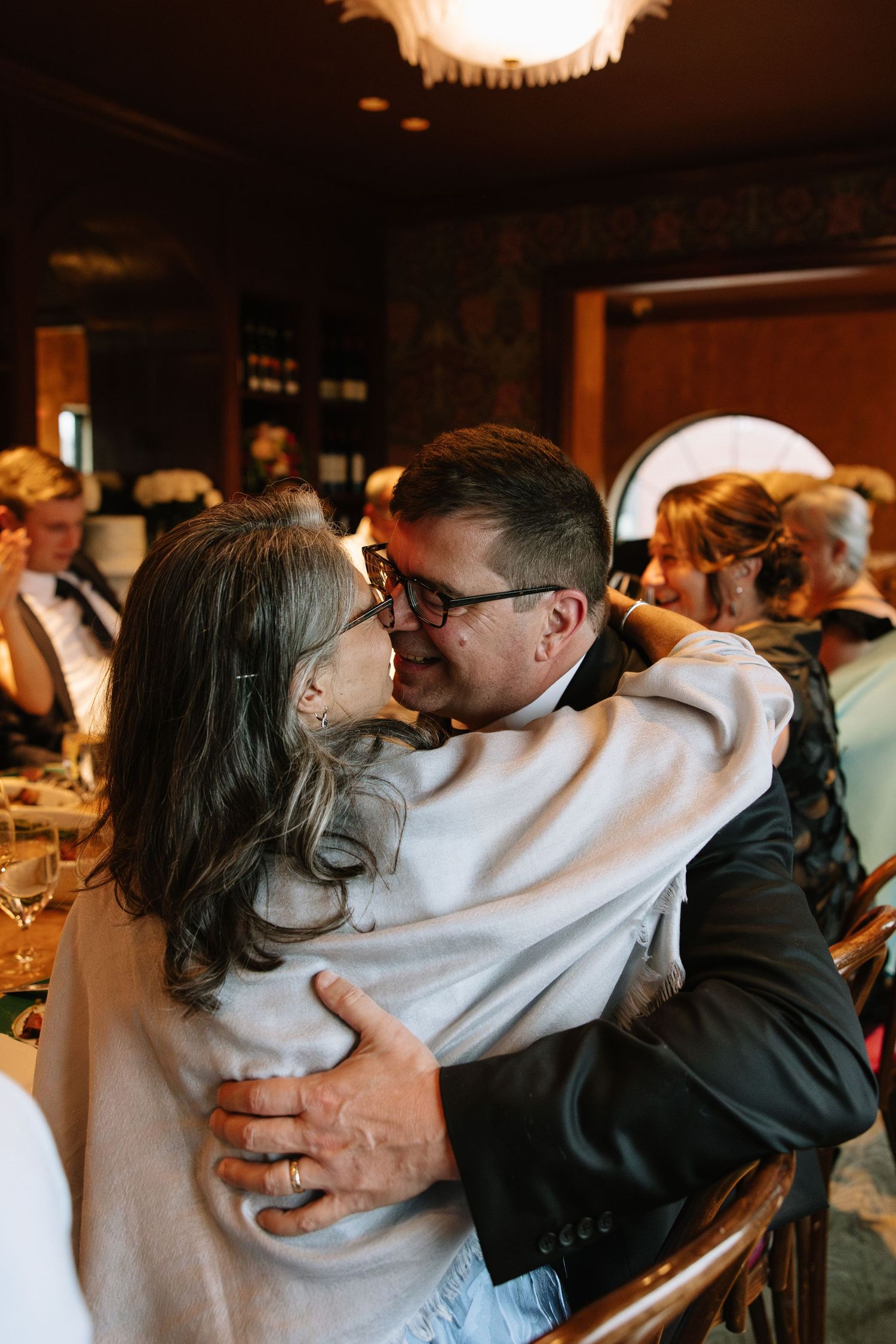 Two wedding guests share a warm embrace and kiss at an elegant indoor wedding reception dinner.