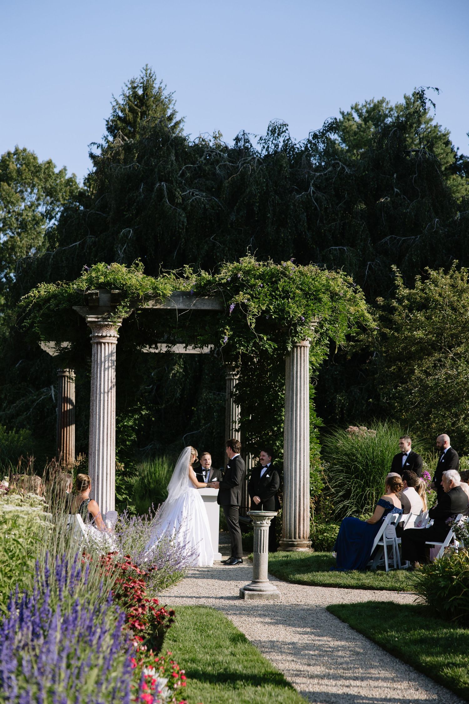 Elegant outdoor wedding ceremony under a vine-covered pergola with stone columns in a lush garden setting.