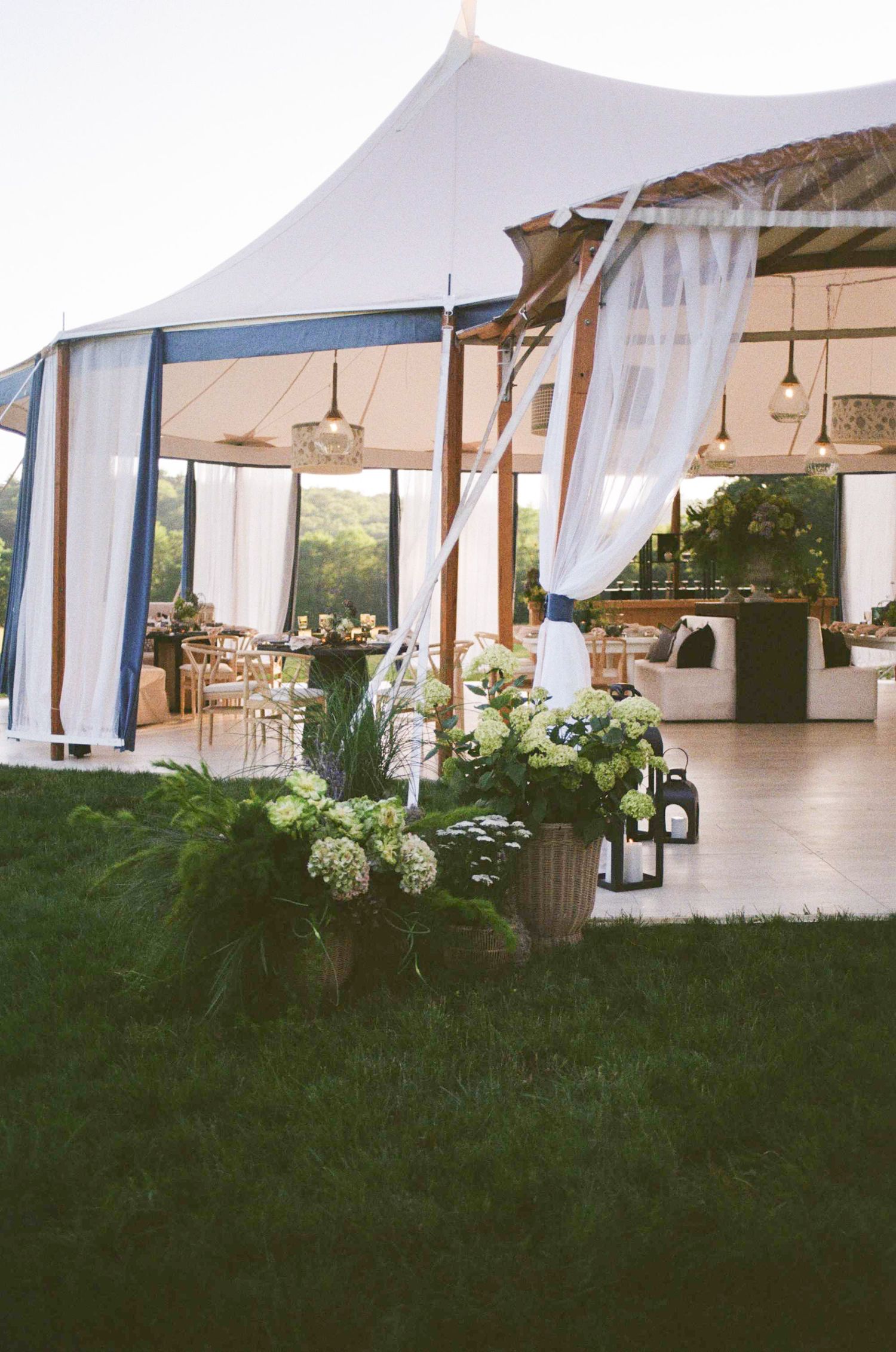 Elegant outdoor wedding tent with white drapes, floral arrangements, and lanterns at dusk.