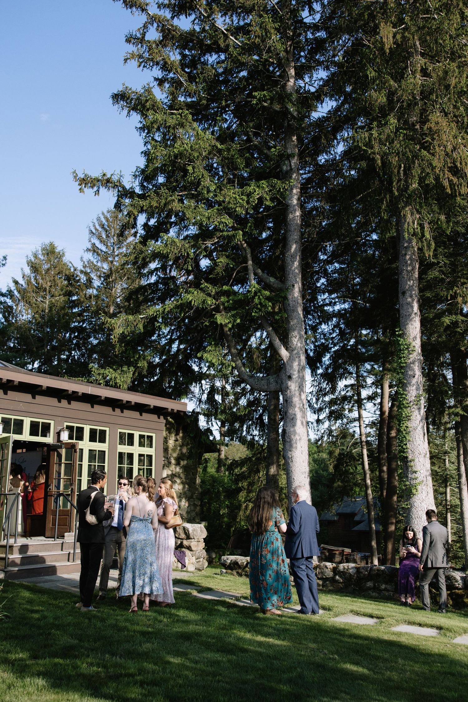 Guests mingle on a green lawn outside a rustic lodge surrounded by tall evergreen trees during an outdoor wedding reception.