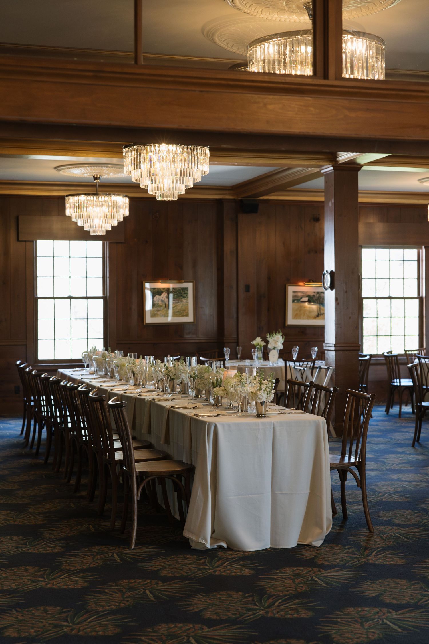 Elegant banquet hall with long white-draped table, crystal chandeliers, dark wood paneling, and floral centerpieces for a formal event.