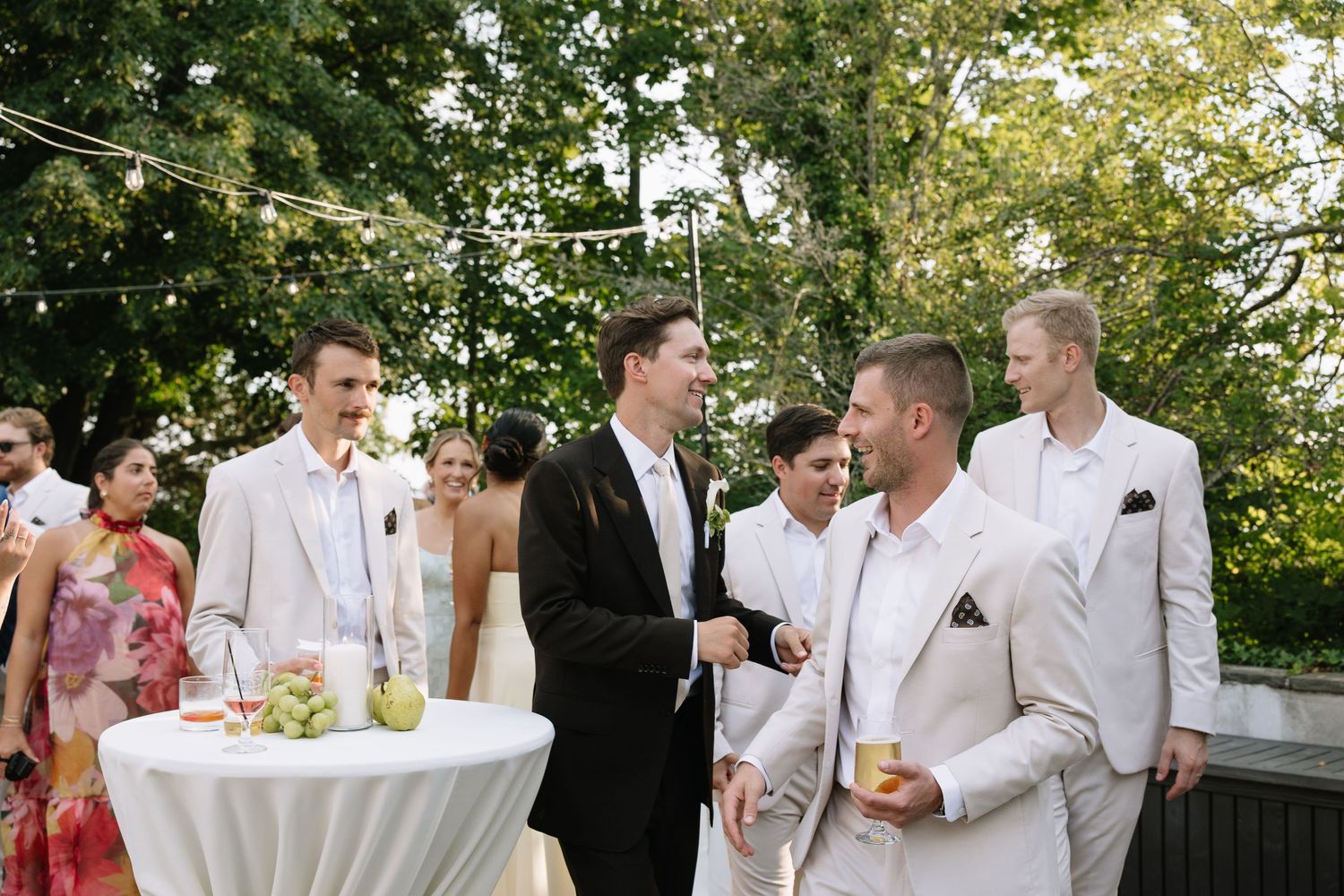 Groomsmen in suits socializing at an outdoor wedding reception with string lights and cocktail tables.