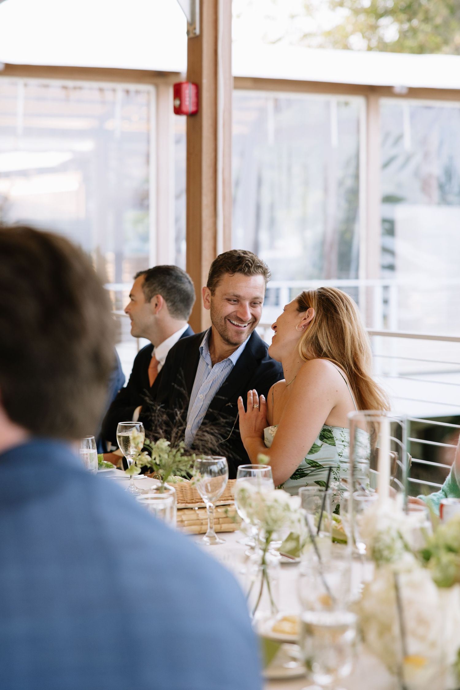 A couple laughing joyfully together at a wedding reception dinner table with floral centerpieces and guests nearby.