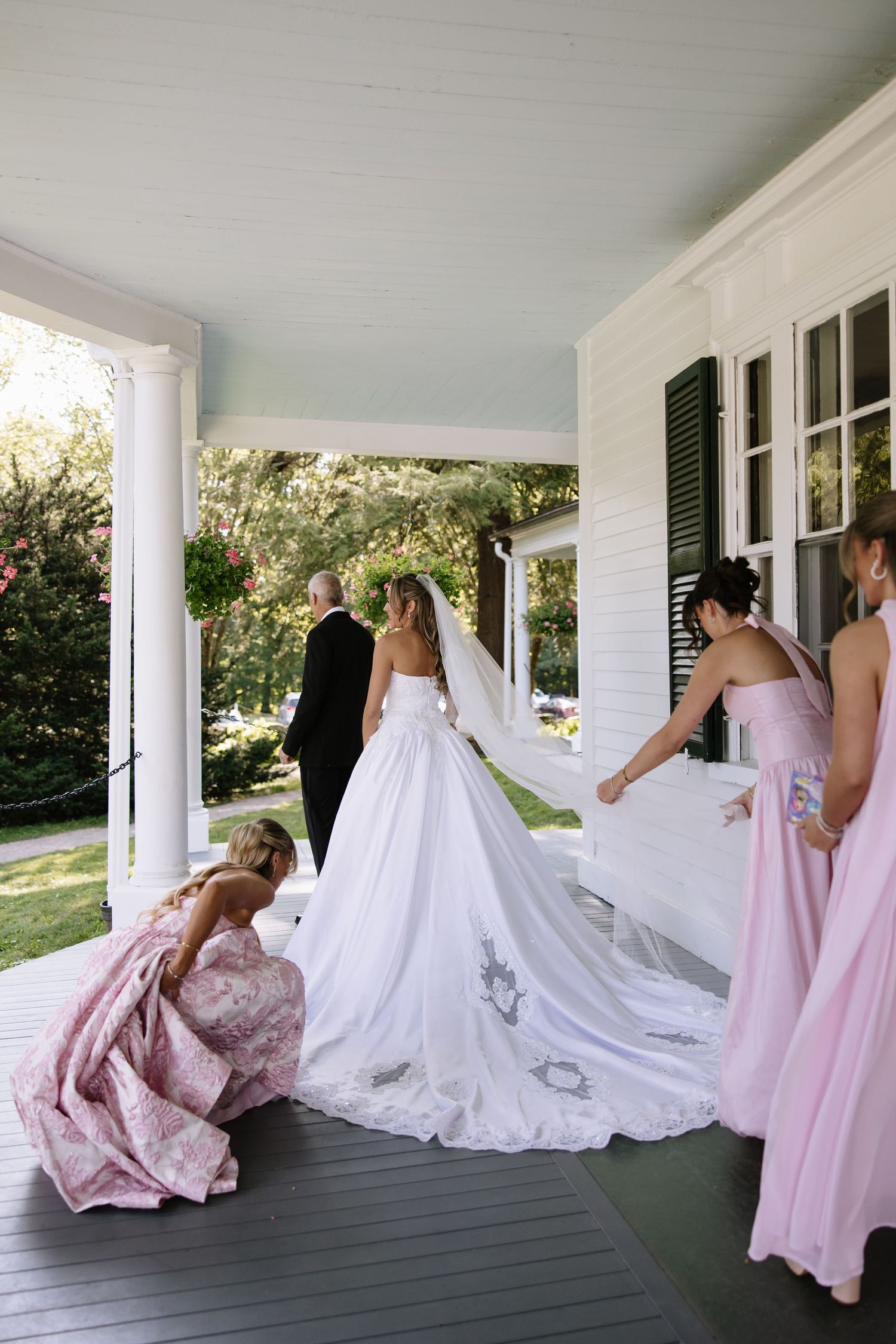 Wedding party in pink bridesmaid dresses and white bridal gown pose on wraparound porch.
