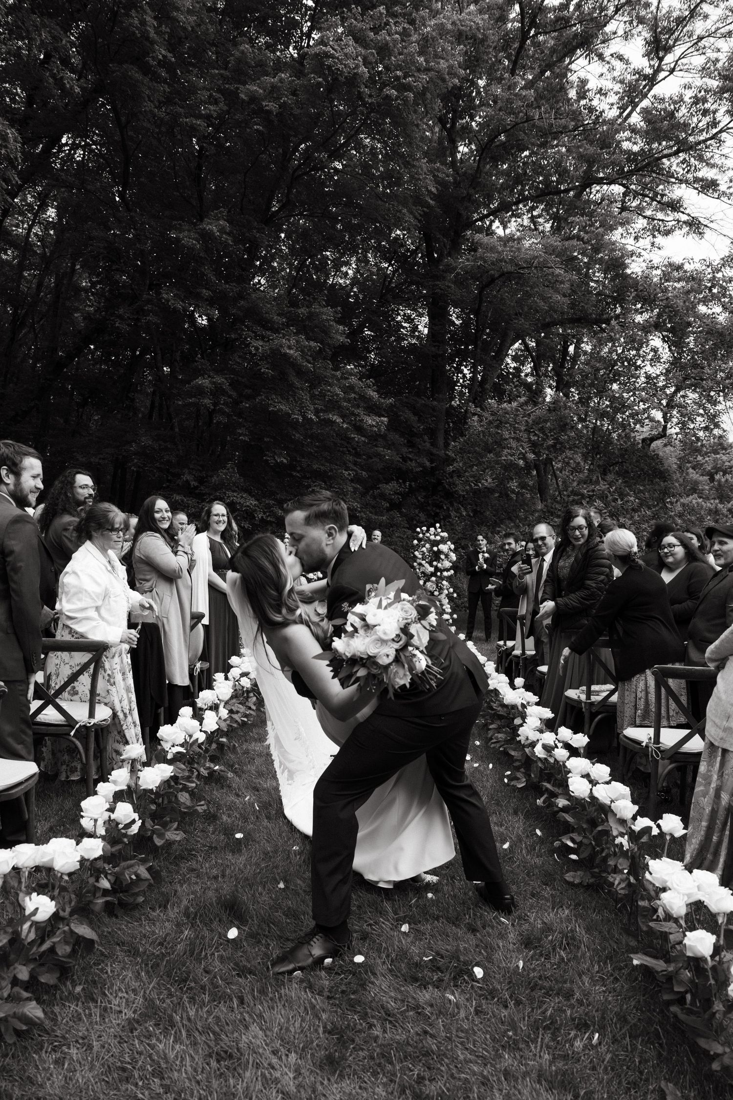 Black and white photo of wedding celebration with guests gathered around floral-lined outdoor ceremony space.