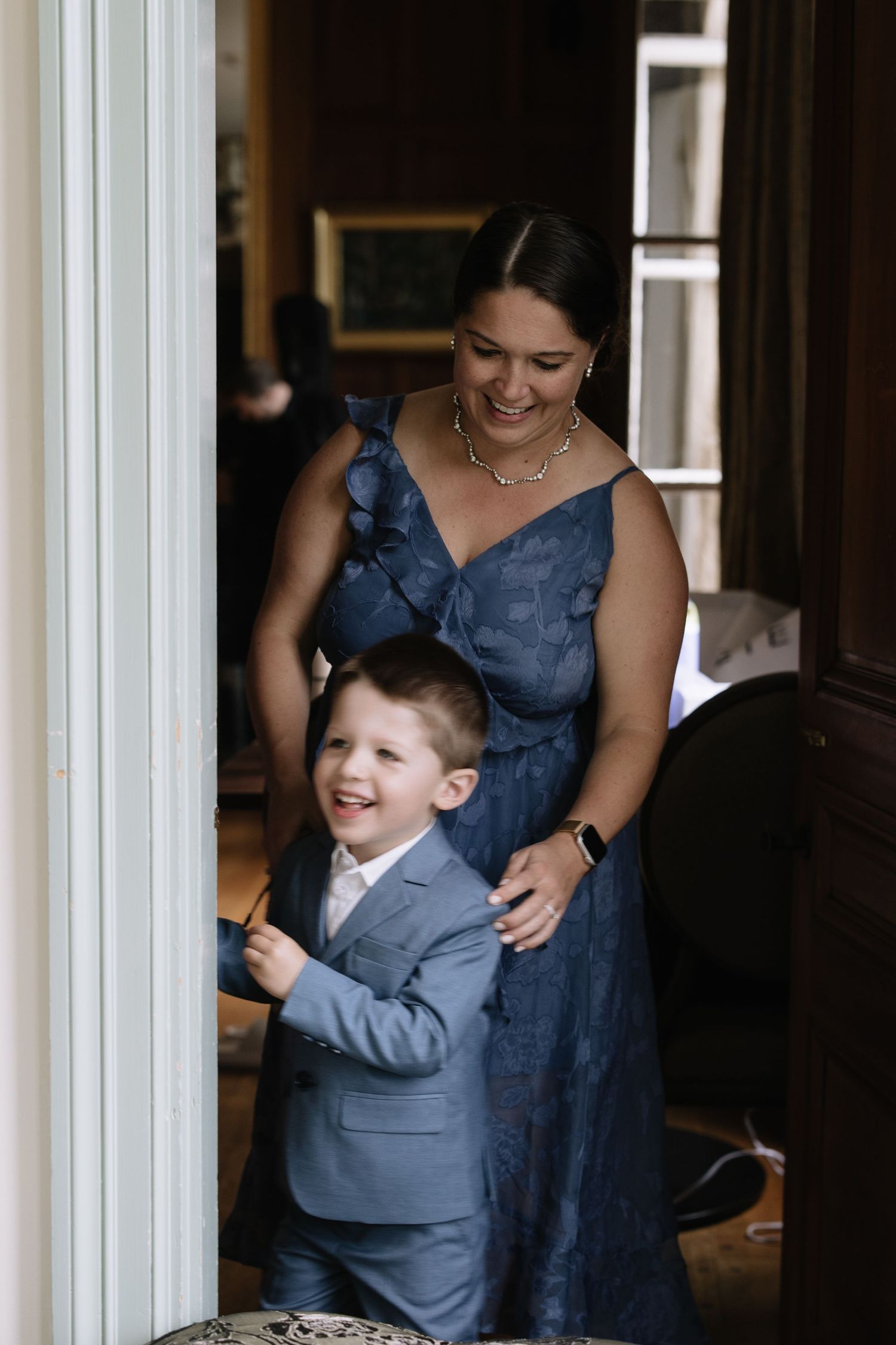 A young ring bearer in a gray suit stands in a doorway with attendants in navy blue dresses.
