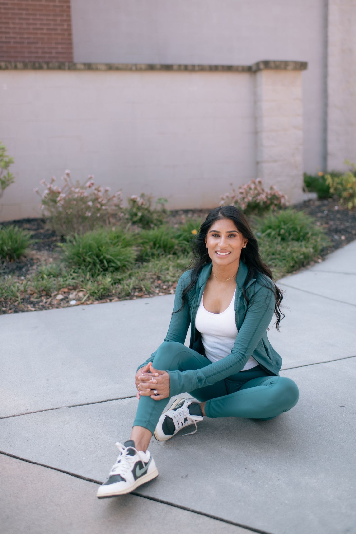 Athletic person in teal workout clothes and sneakers stretching on concrete sidewalk near brick wall.
