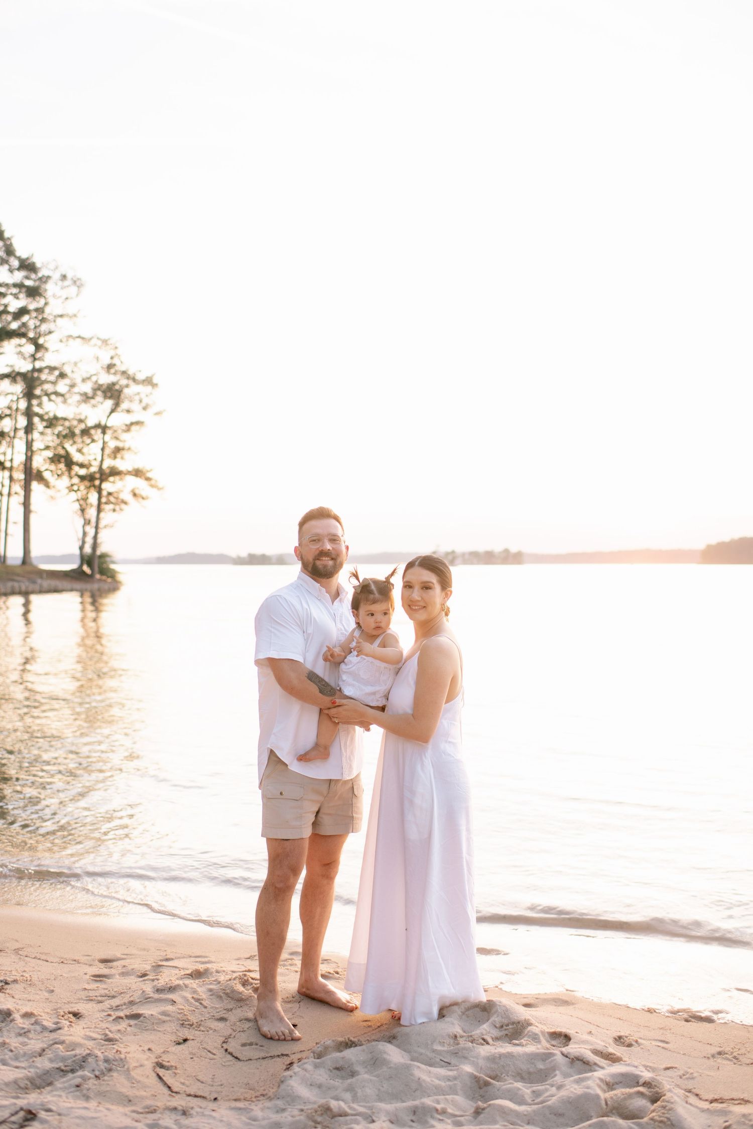 Family in white outfits posing together on sandy beach with water and trees in background.
