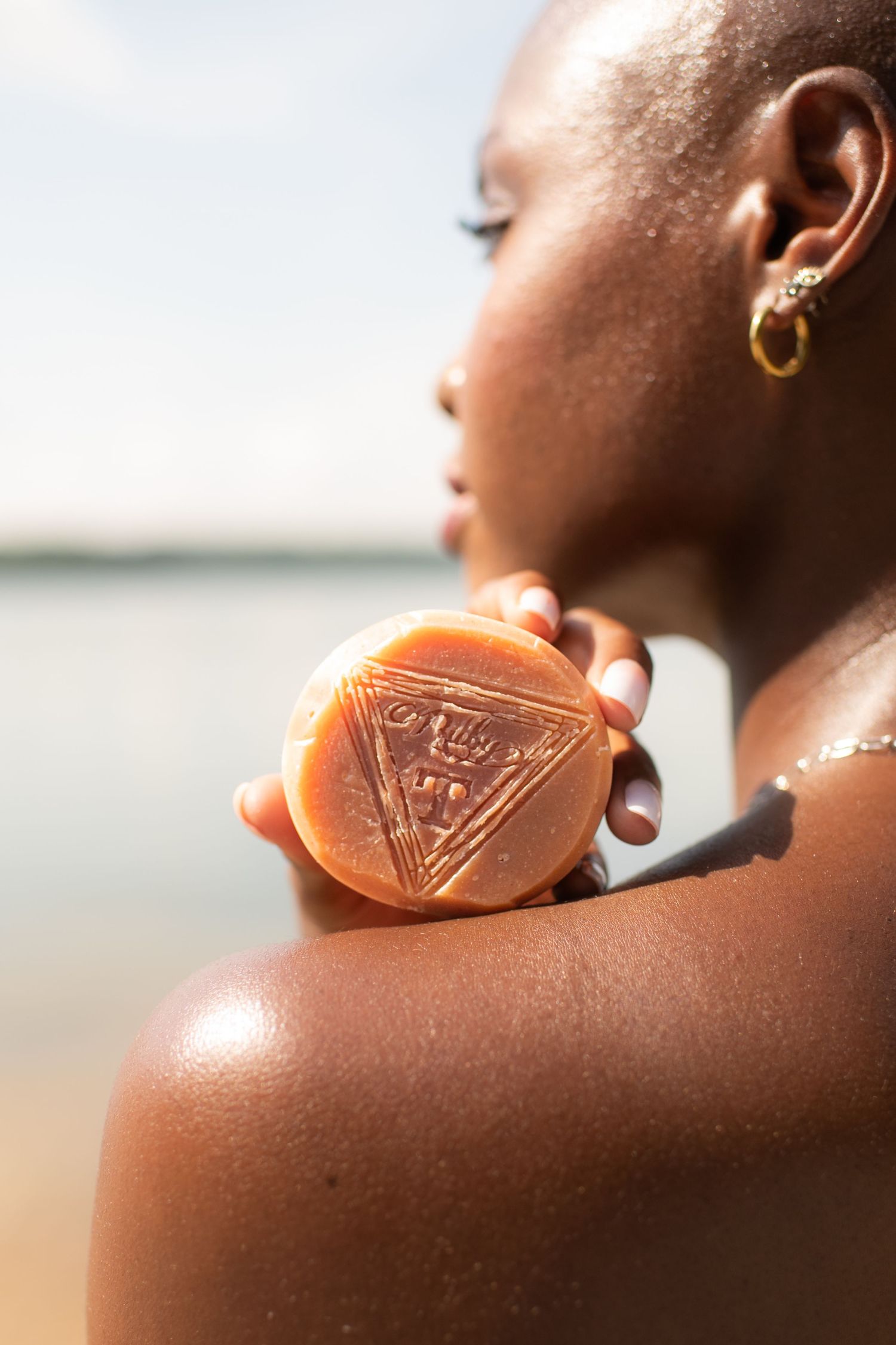 Woman applying an orange soap bar to her shoulder near a beach, wearing gold earrings.