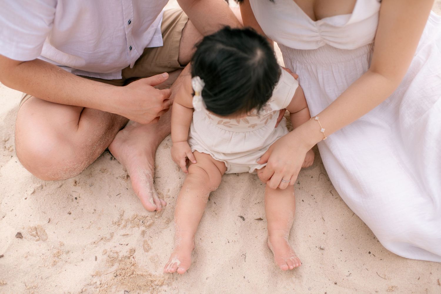 A small baby wearing a white outfit sits on sand between two people in light colored clothing.