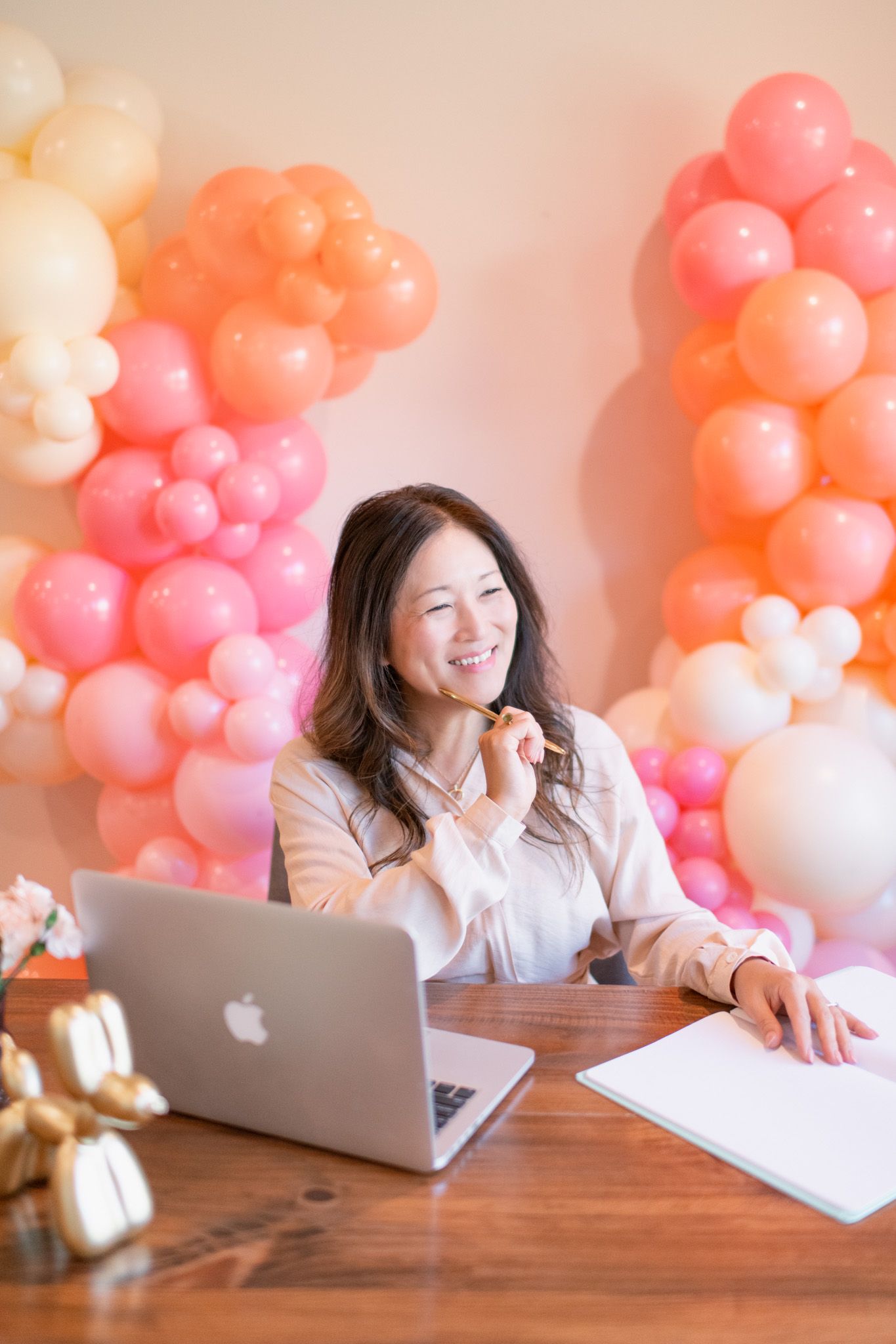 A workspace decorated with pink and coral balloon garlands featuring a laptop and writing materials on a wooden desk.