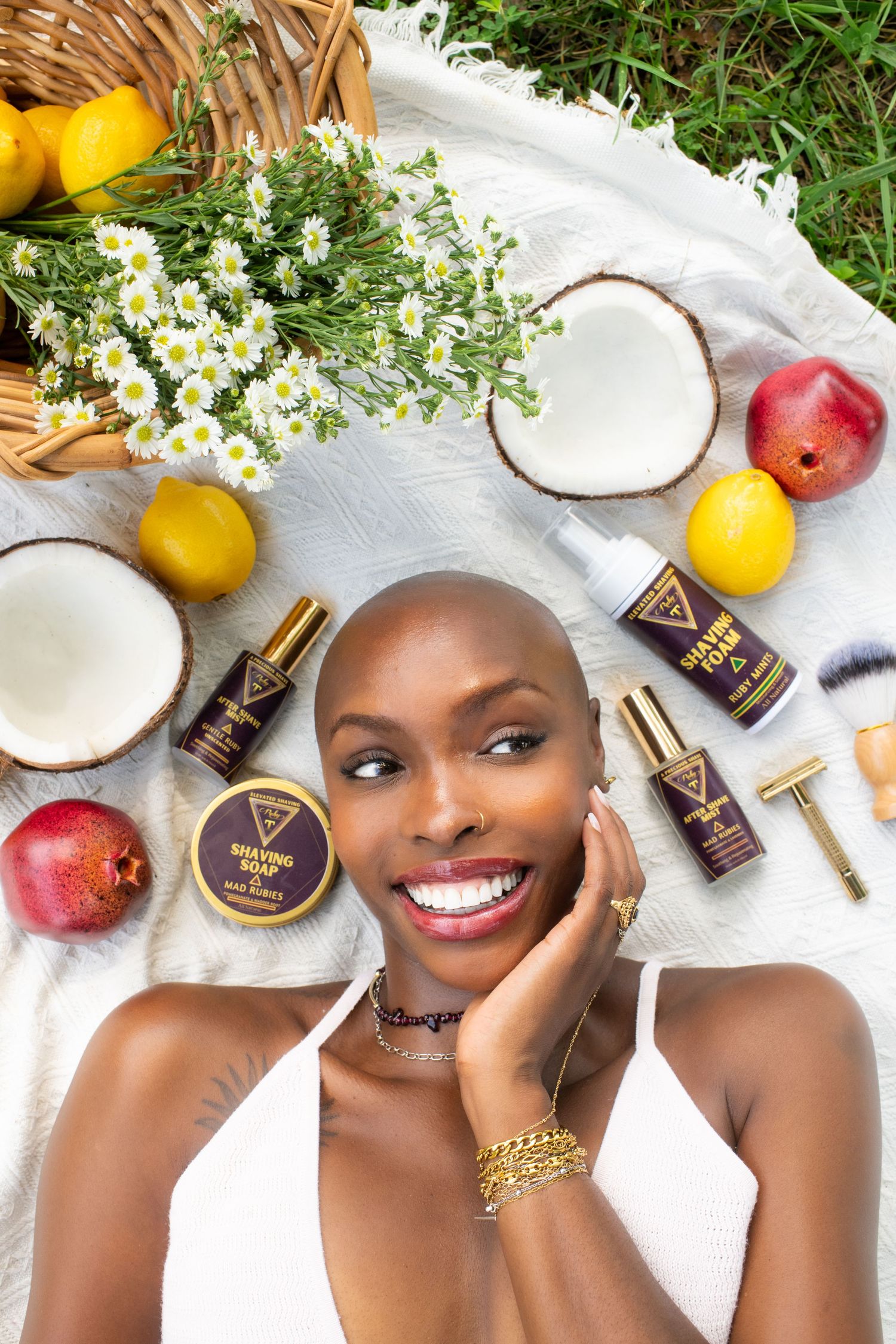 Smiling woman with shaved head lying among skincare products, fruits, coconut, and daisies on a white blanket outdoors.