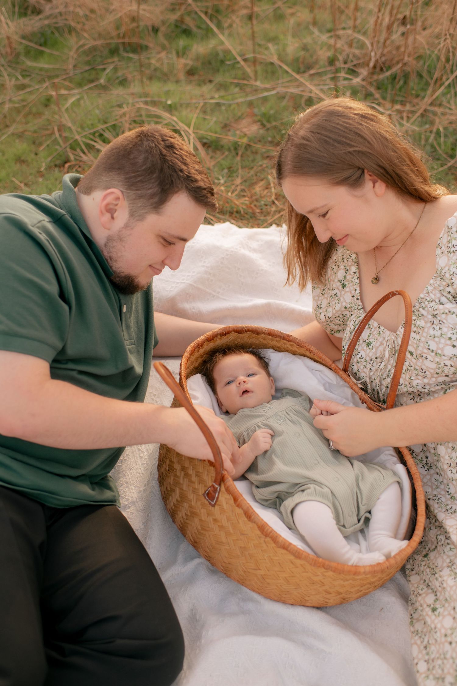 A couple sits on a blanket outdoors looking lovingly at their baby resting in a woven picnic basket.