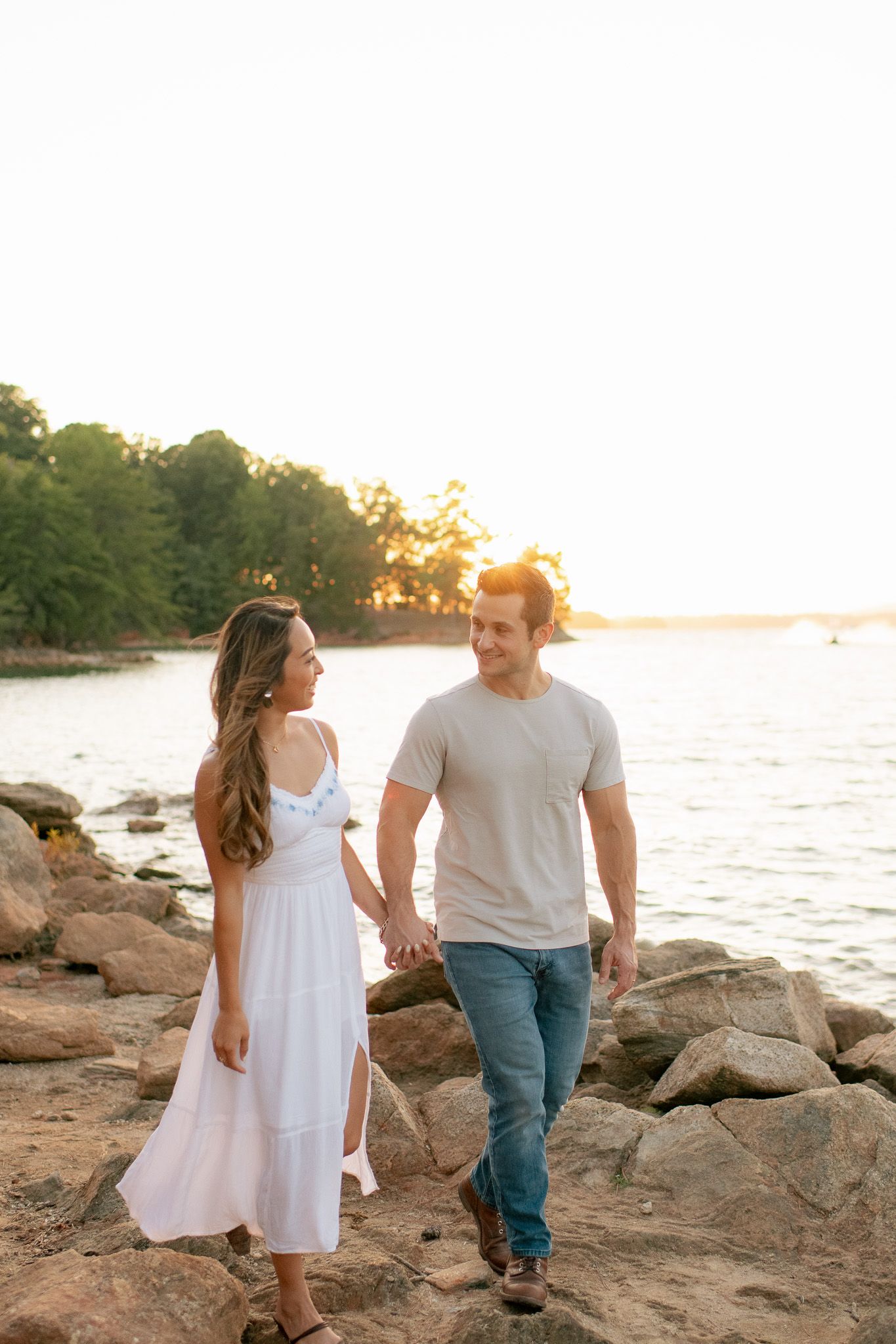 Two people hold hands walking along a rocky beach shore during golden hour sunset.