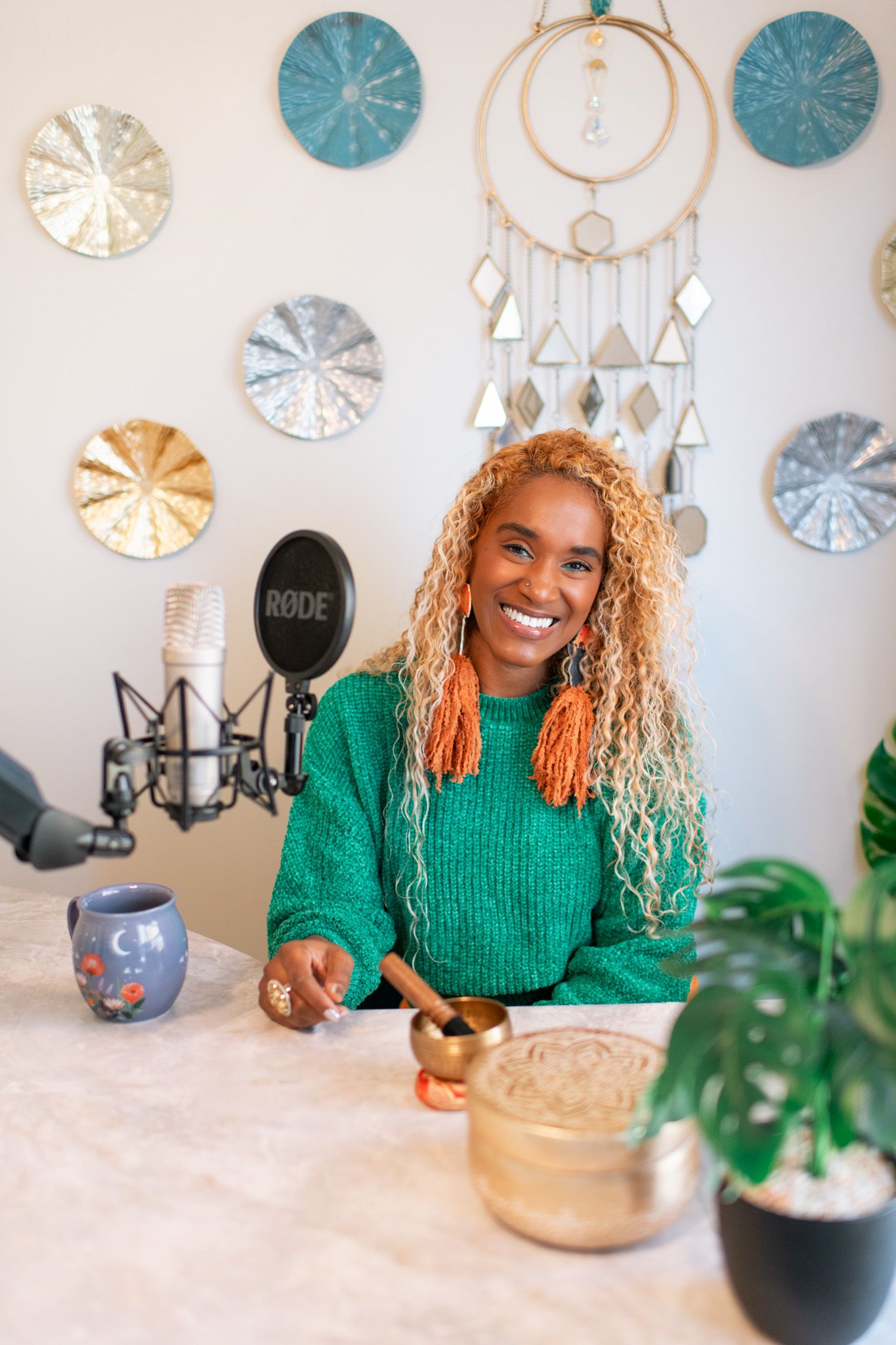 A person in a green sweater sits at a desk with microphones and decorative wall art in a podcast studio setup.